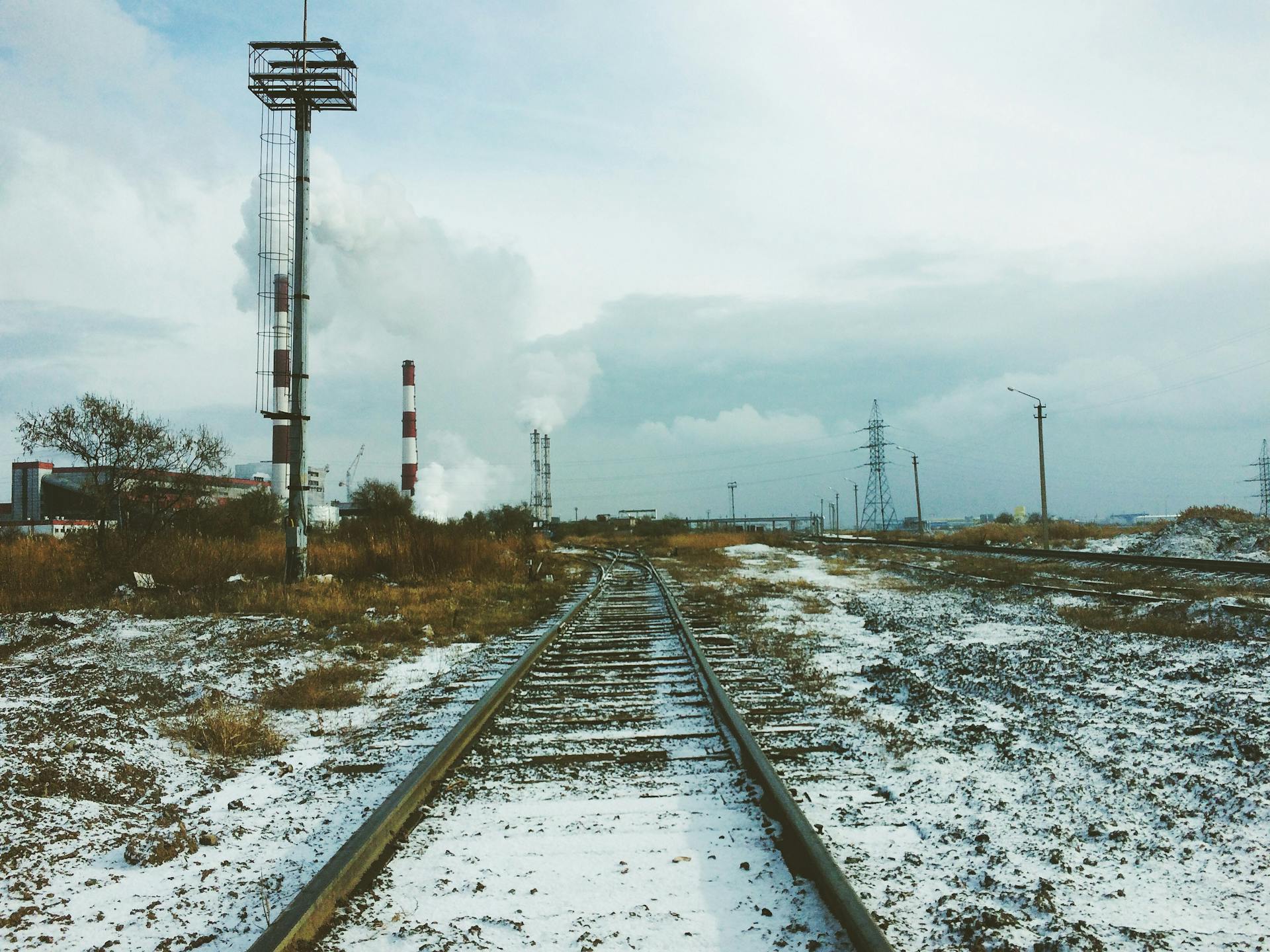 Abandoned railway tracks in winter with industrial smokestacks and utility poles under a cloudy sky.