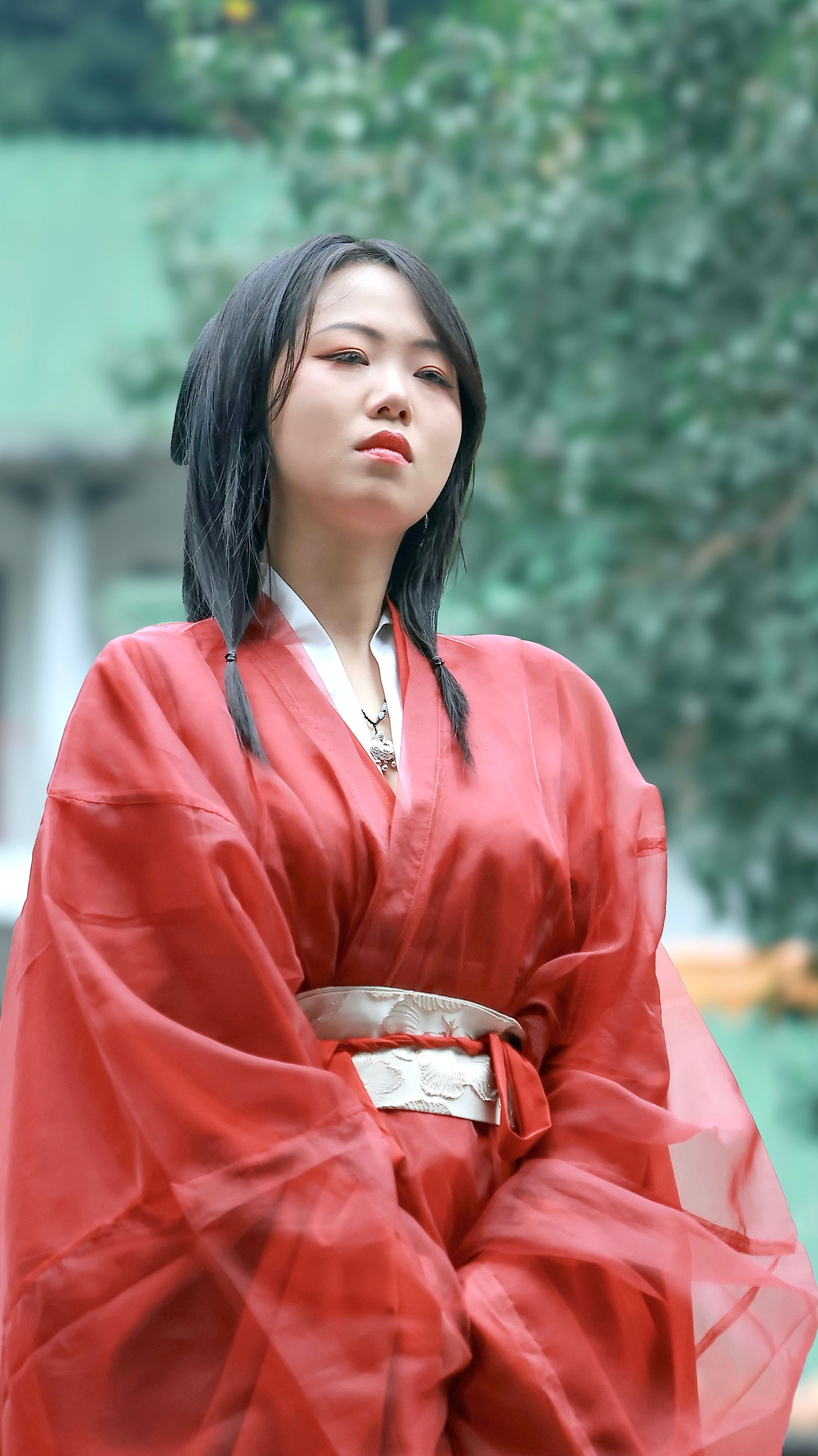 Asian woman in a red traditional robe posing outdoors, serene expression.