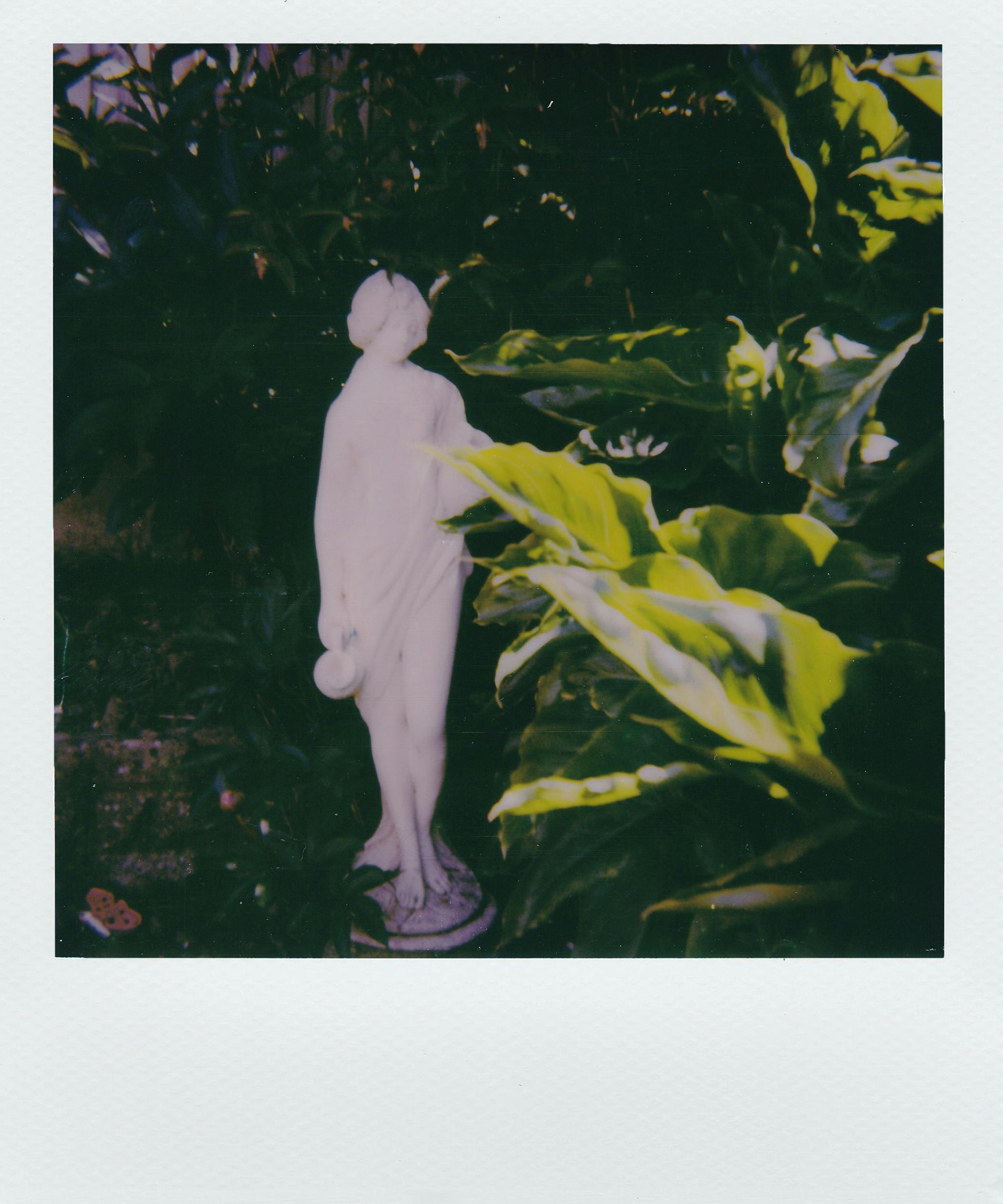 A Greek-style statue amidst lush greenery, captured on instant film for a vintage feel.