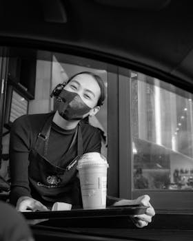 Barista wearing a mask serves coffee through a drive-thru window with a friendly smile.