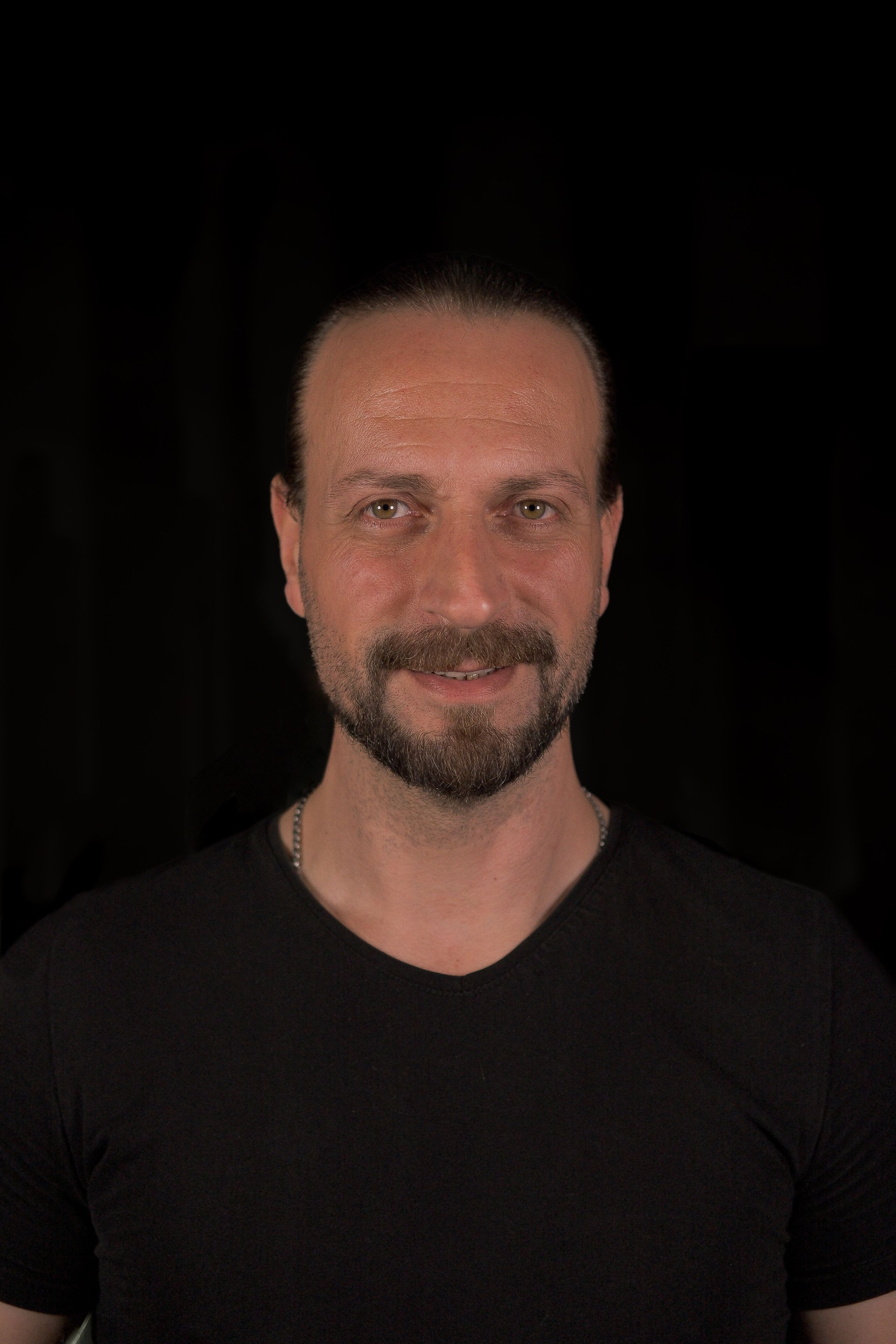 Studio portrait of an adult man smiling against a black background, wearing a black shirt.