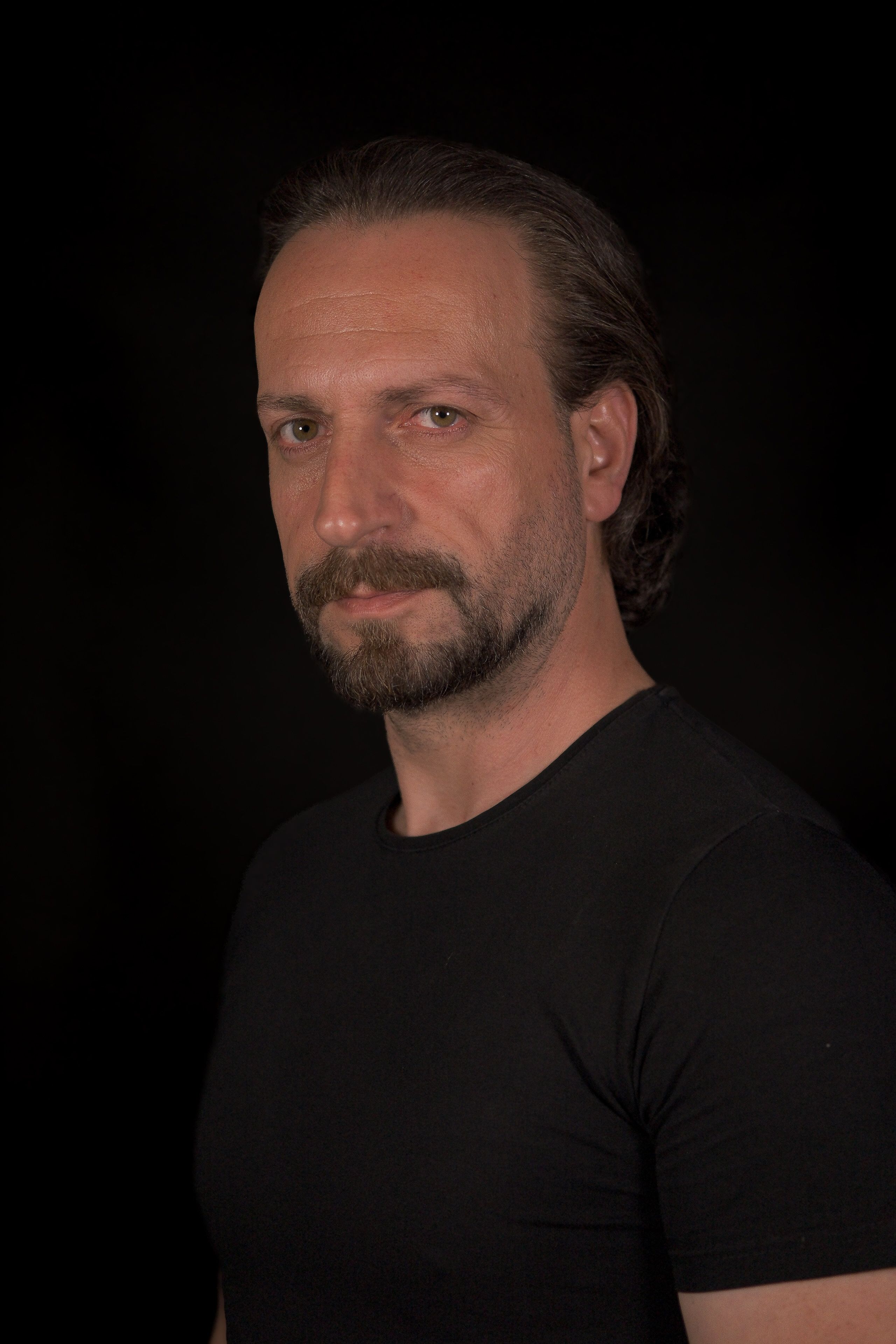 Studio portrait of a confident bearded man in a black shirt against a dark background.