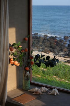 Calming ocean view through window with vase of oranges, shells.