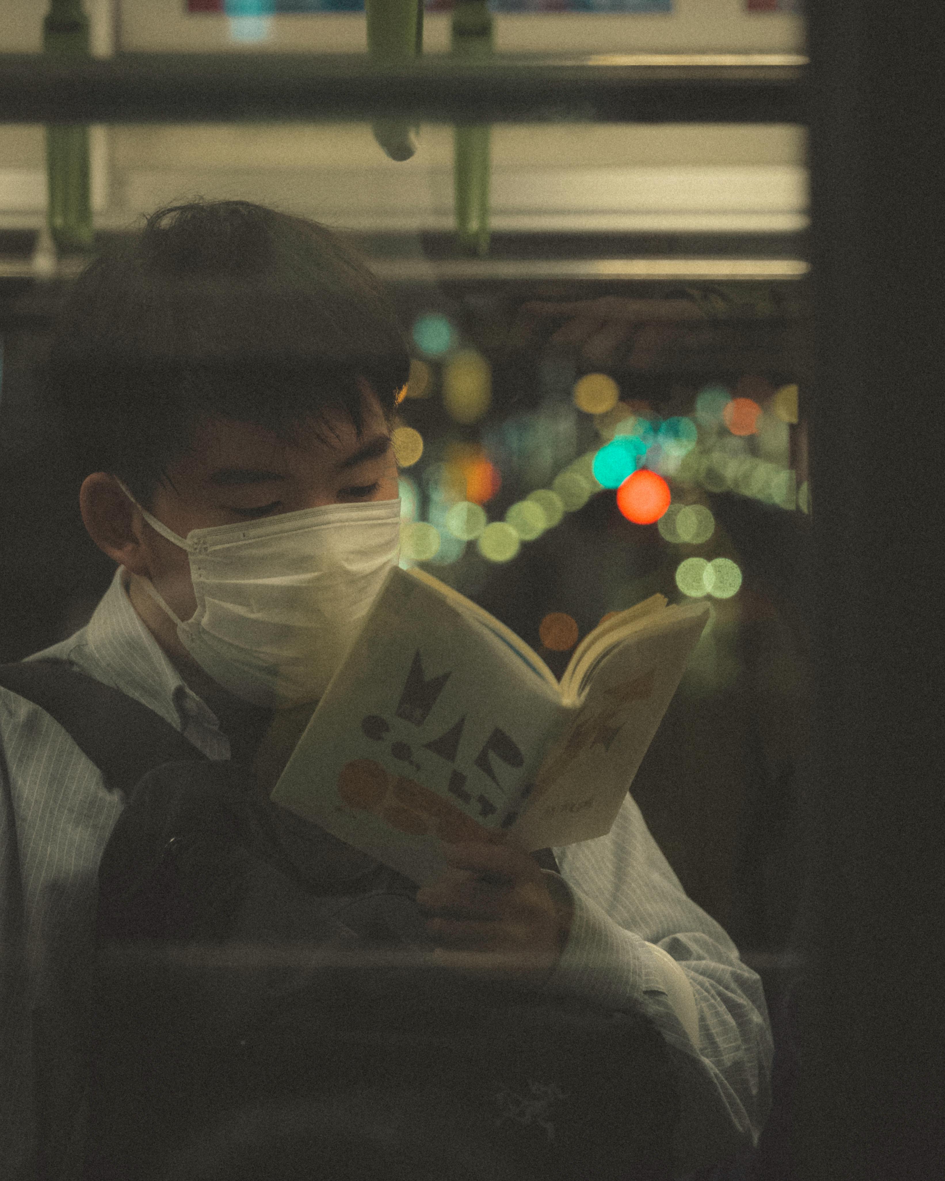 Young adult reads a book wearing a mask on Tokyo subway. City lights blur in the background.