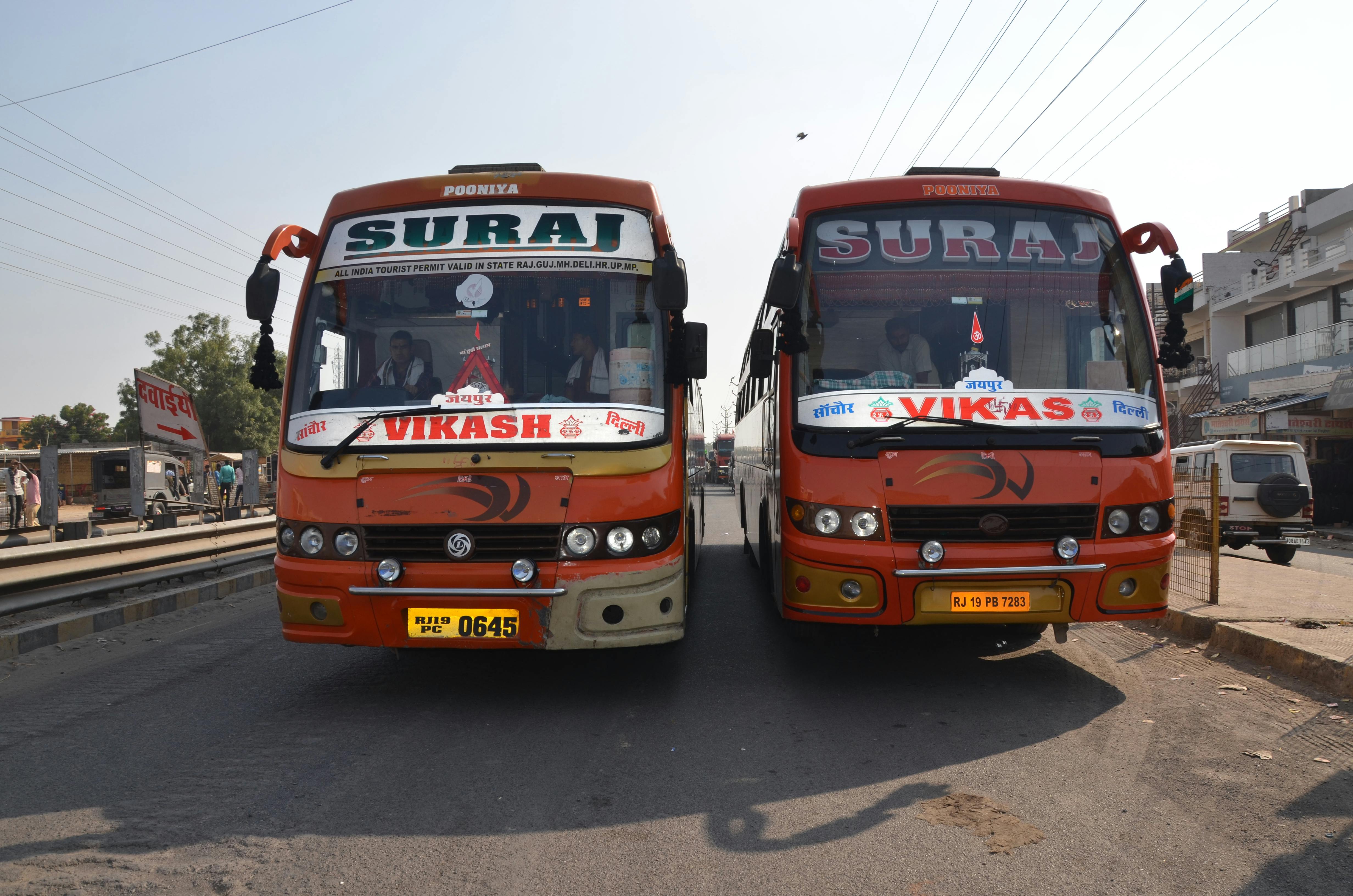 Two vibrant Indian buses parked on a city street in broad daylight with urban surroundings.