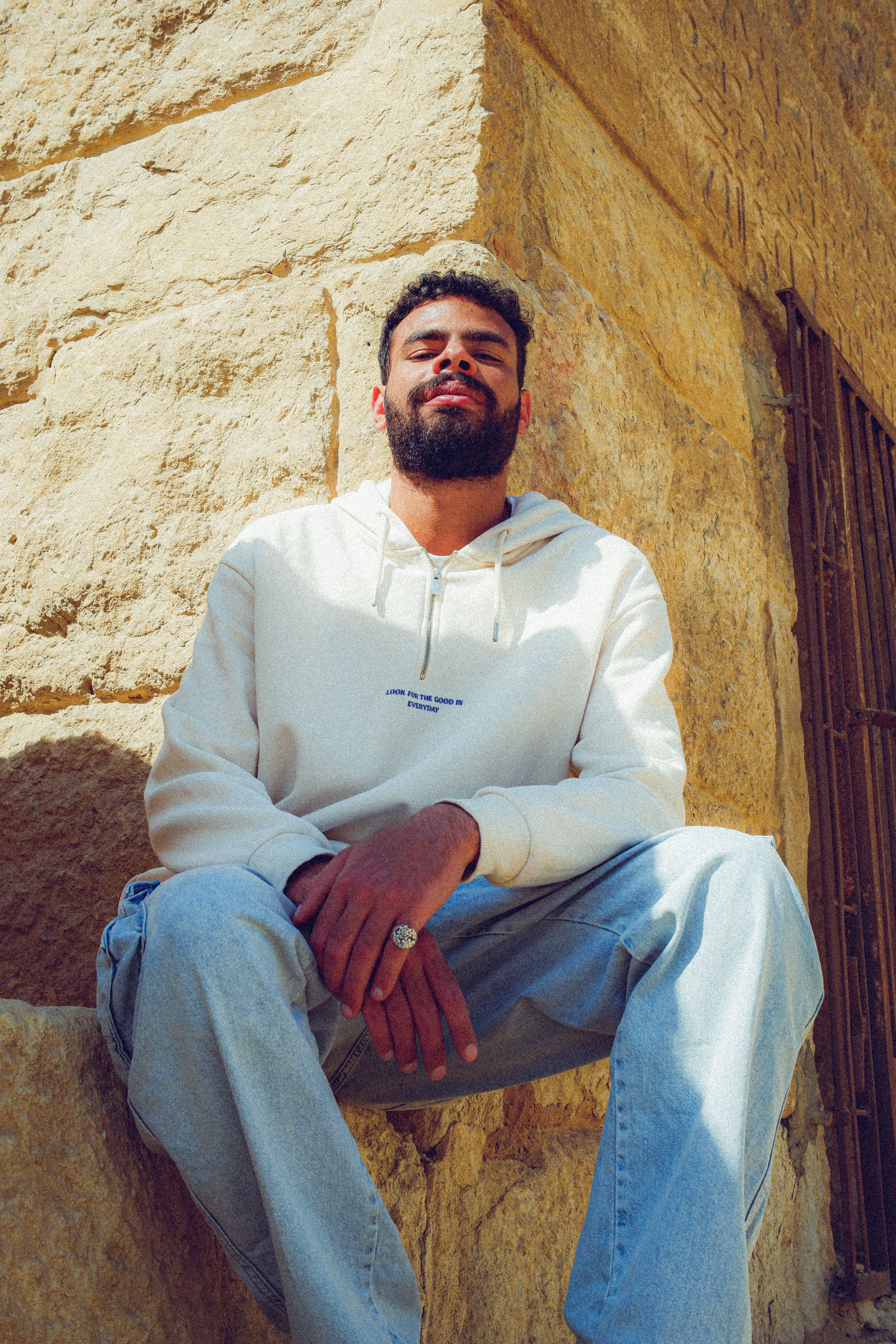 Man in casual outfit sitting against a stone wall enjoying sunlight.