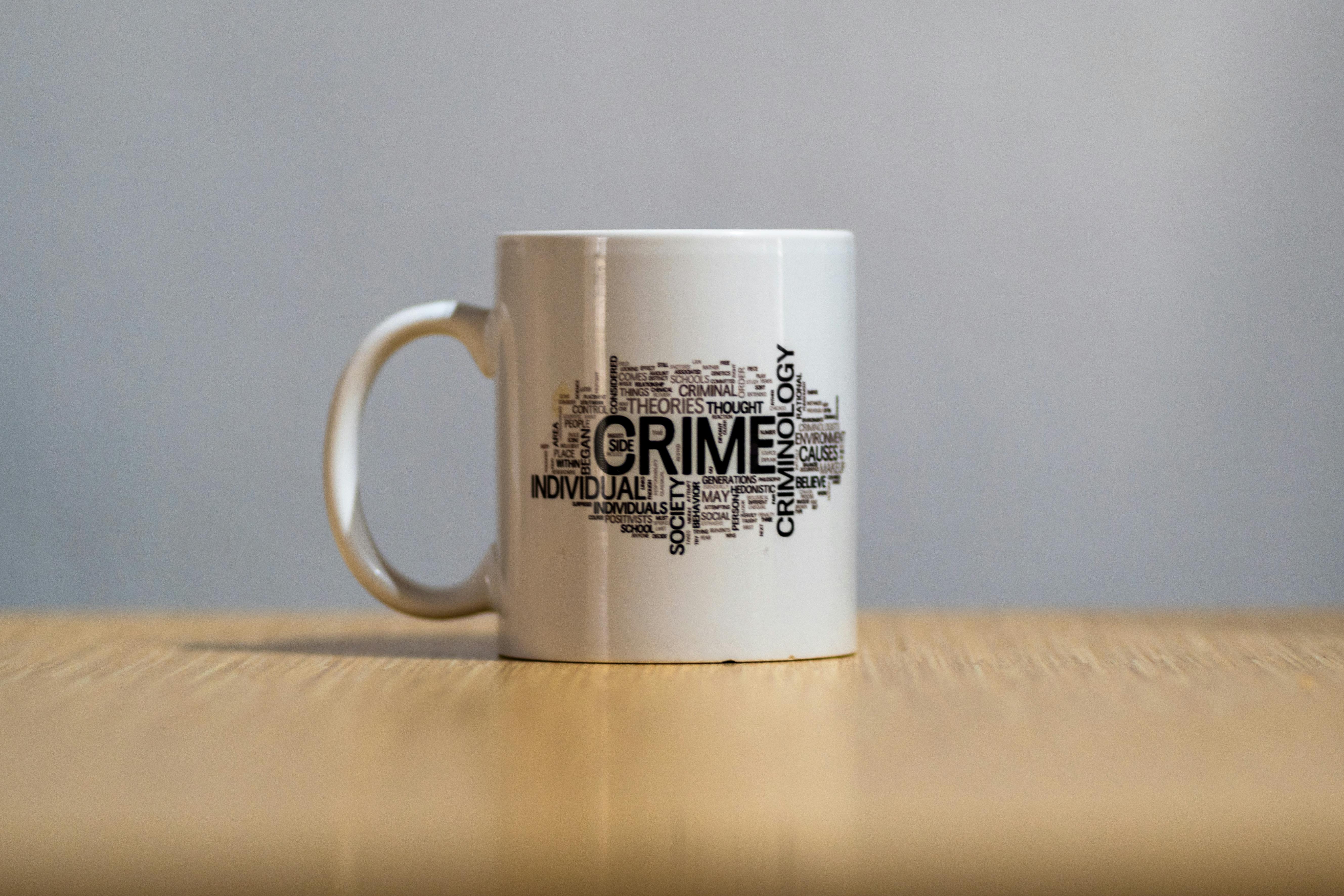 A white coffee mug on wooden surface with a unique crime-themed text design.