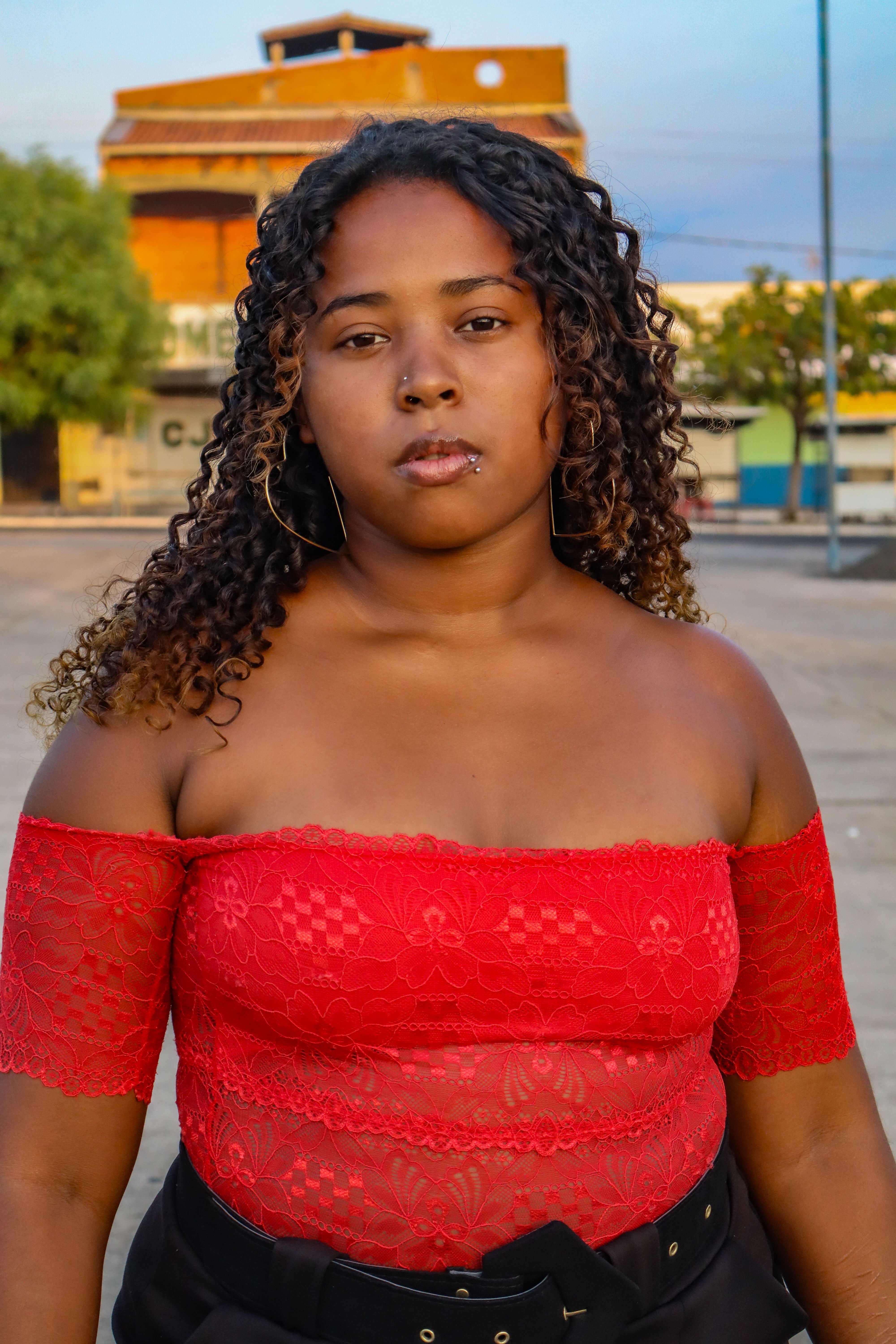 Confident young woman in lace red top posing outdoors with urban backdrop.