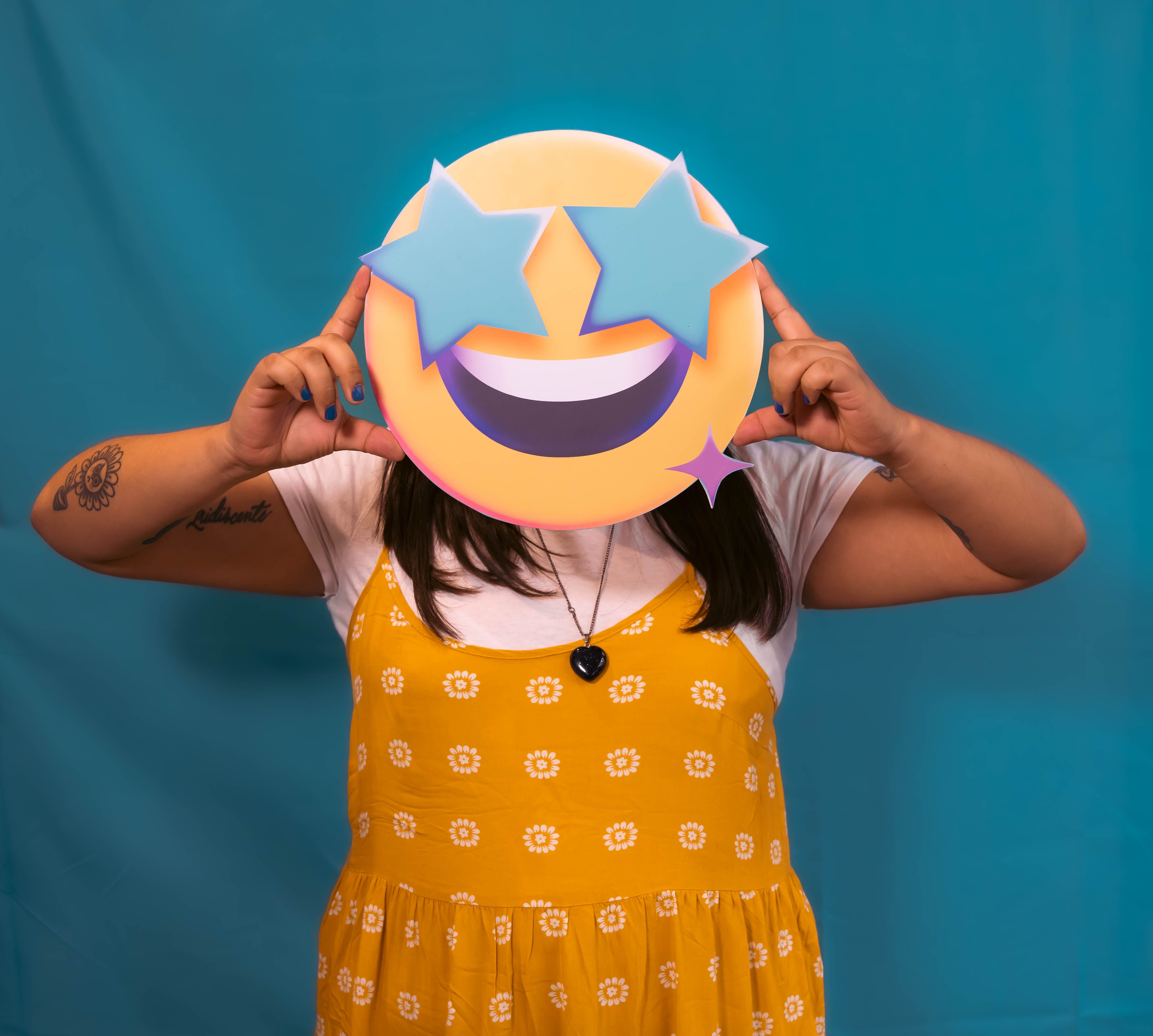 Person holding a star eyes emoji mask over face in a playful pose, wearing a yellow dress.