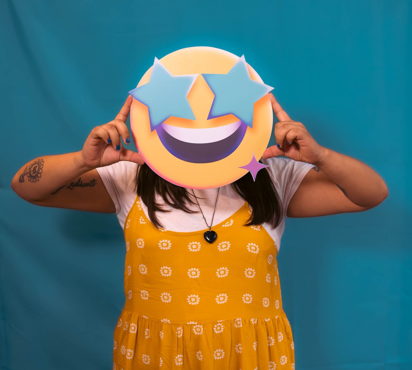 Person holding a star eyes emoji mask over face in a playful pose, wearing a yellow dress.