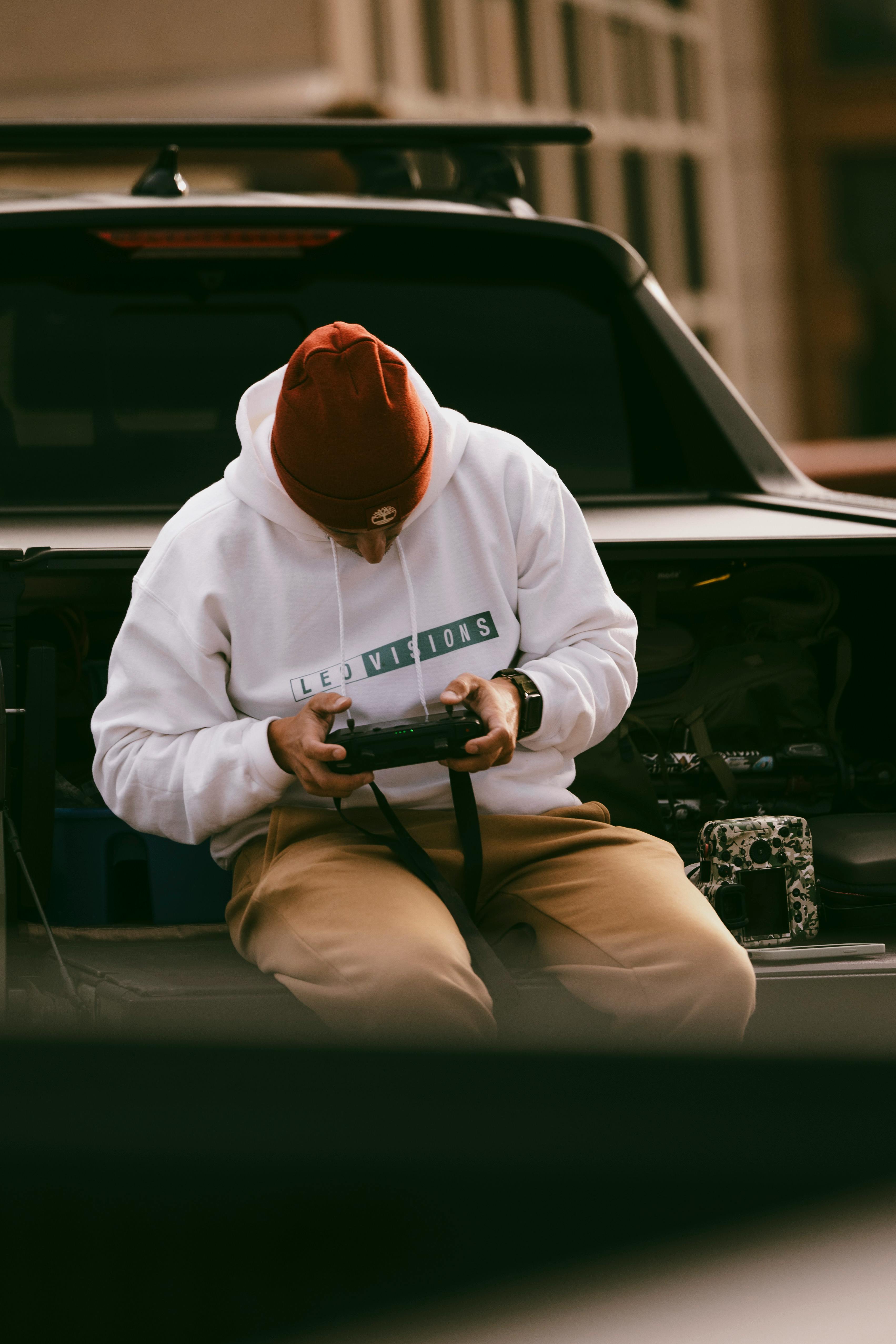 A man in a hoodie and beanie sits on a car trunk controlling a drone remote.