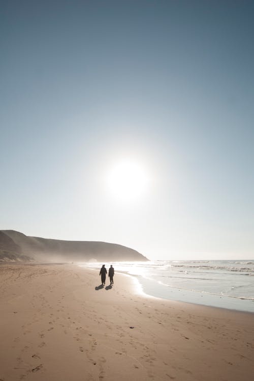 Silhouettes of unrecognizable travelers strolling slowly on empty sandy coastline with waving sea on sunny summer day