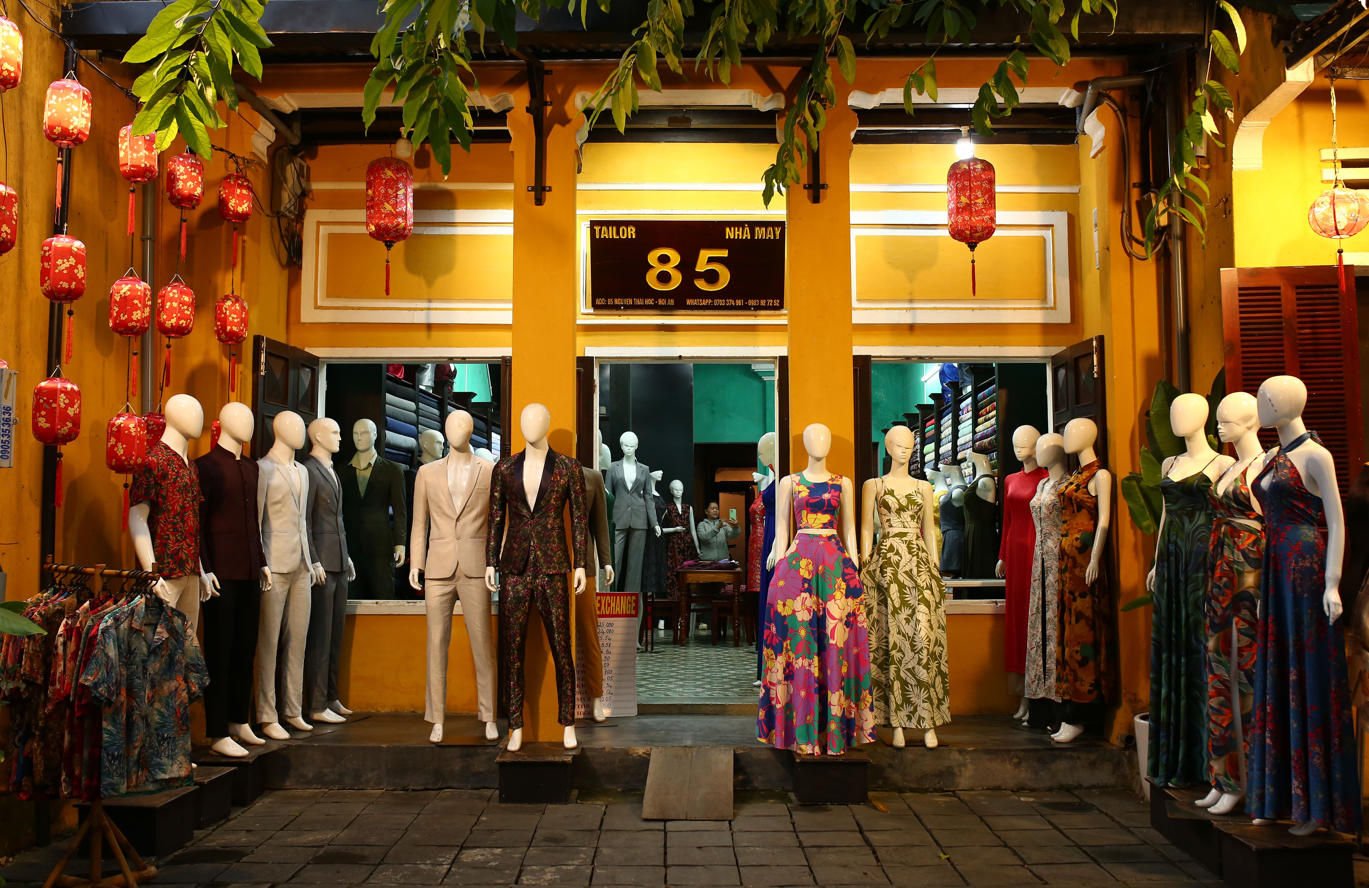 Mannequins display colorful attire outside a Hội An tailor shop at night, illuminated by lanterns. Mannequins display colorful attire outside a Hội An tailor shop at night, illuminated by lanterns.