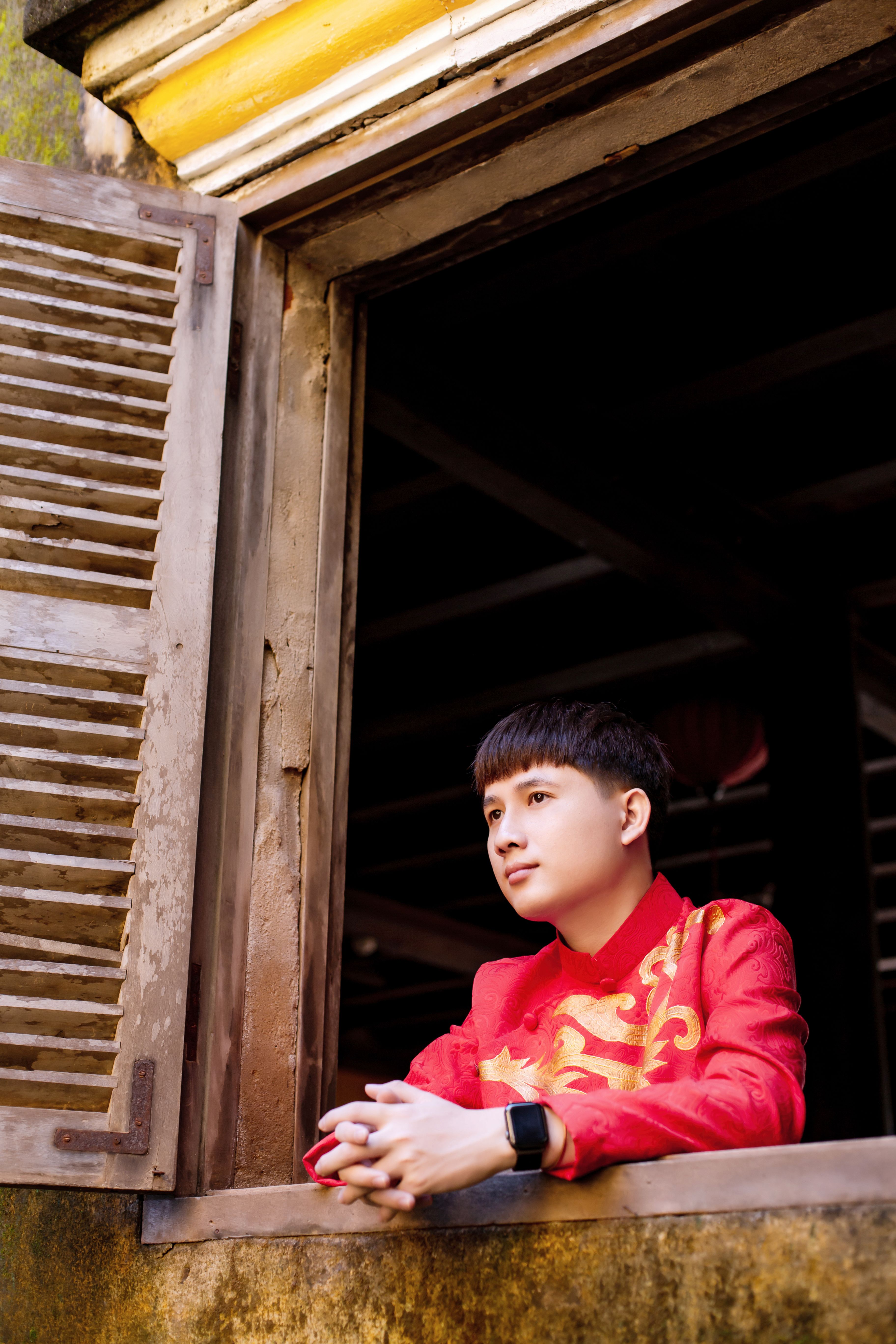 A young man in vibrant red áo dài leaning on a vintage window in Hội An, Vietnam.