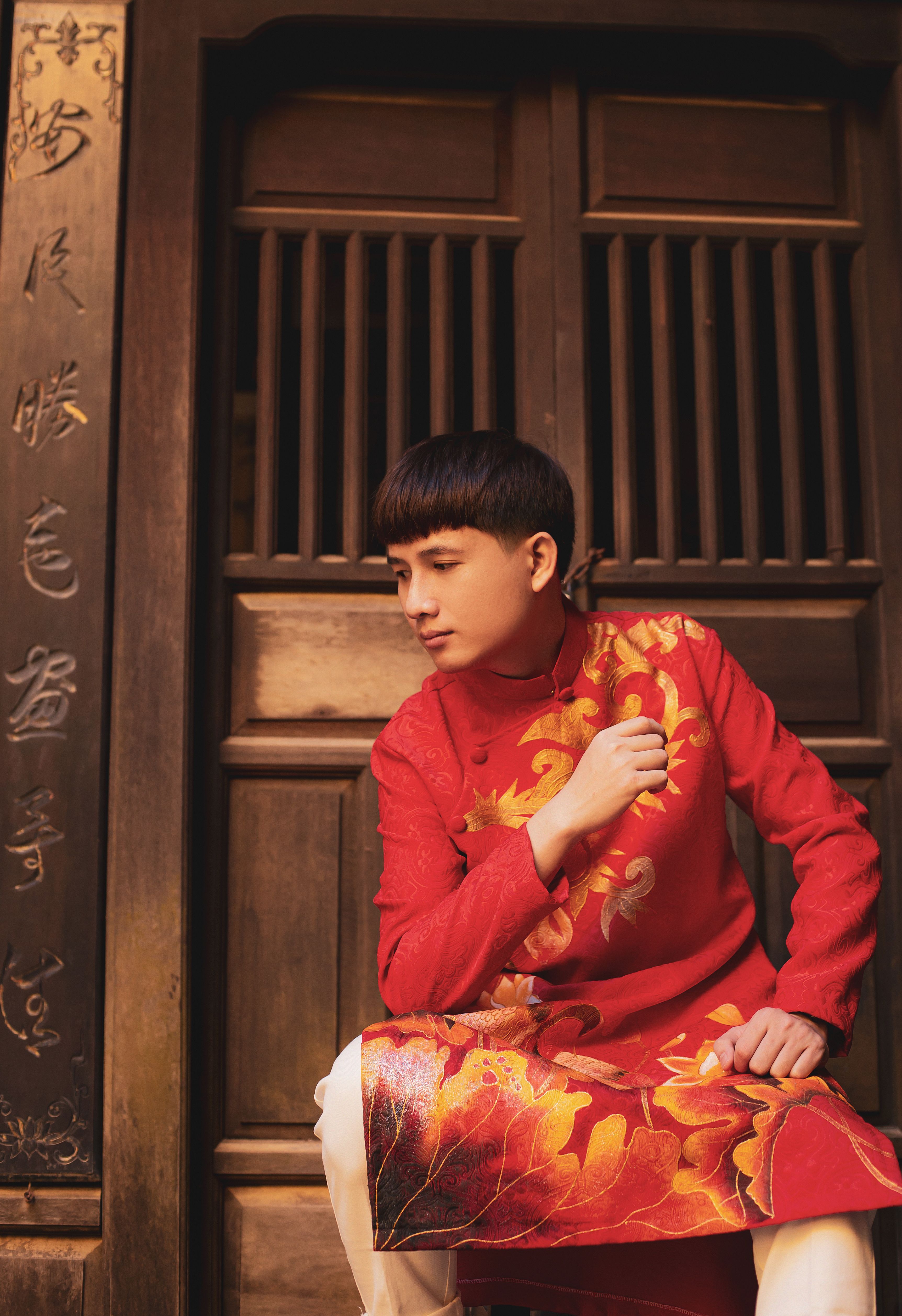 Man in traditional red Vietnamese dress posing by a wooden door in Hội An.