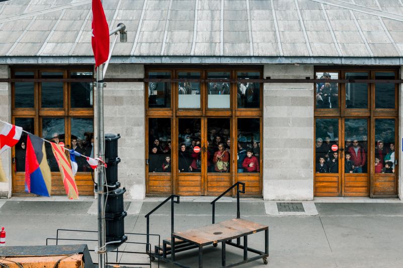 A diverse group of people looking out through large windows in an urban setting with colorful flags outside.