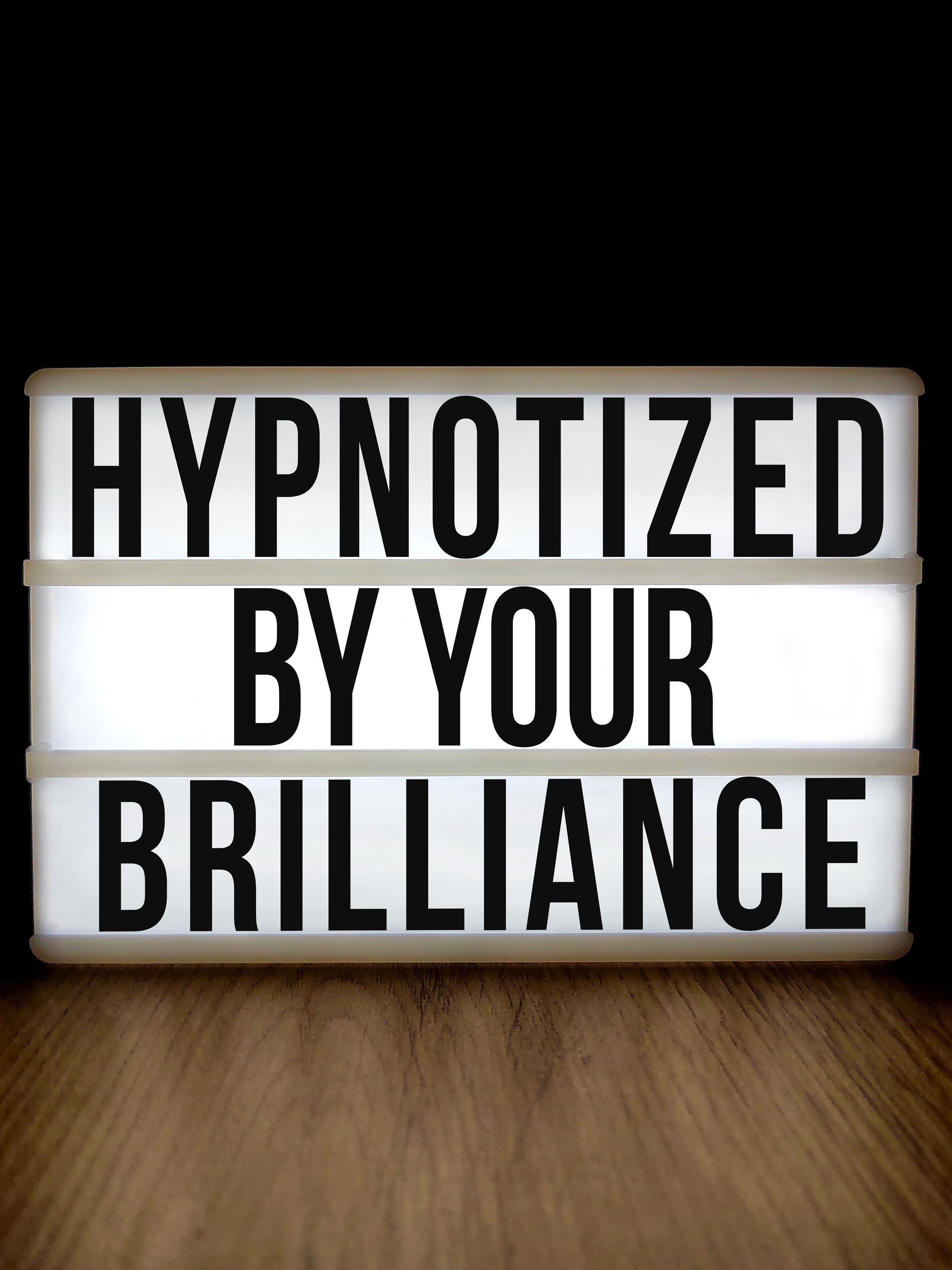 Lightbox with an inspirational message 'Hypnotized by Your Brilliance' on a wooden surface.