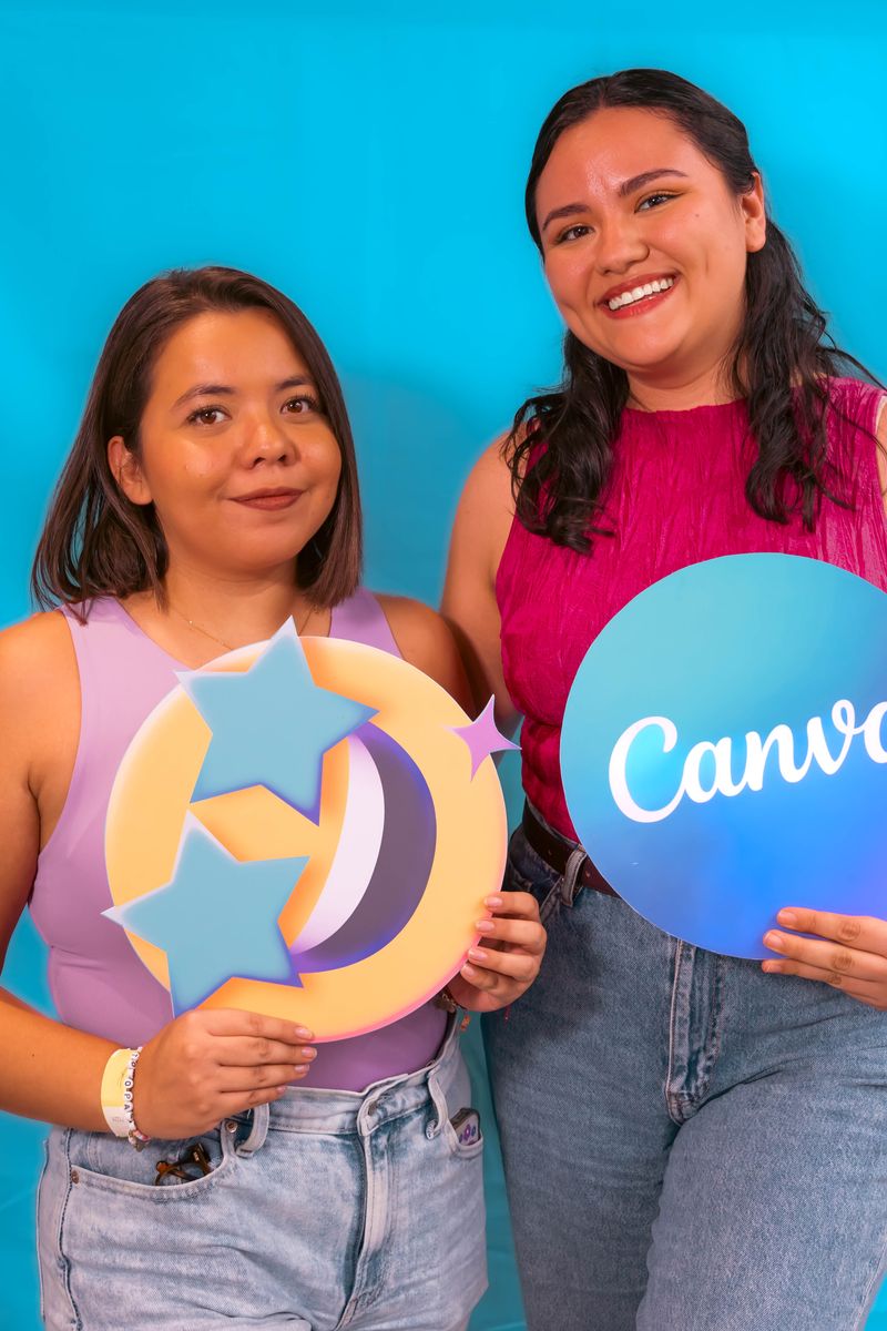 Two women smiling and holding a Canva sign against a blue background, promoting creativity and design.