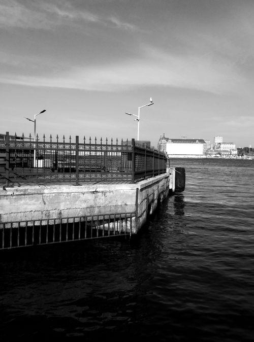 Free A black and white image of a waterfront dock with city buildings in the background. Stock Photo