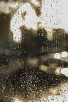 Close-up of raindrops on a window with blurred city lights at dusk, creating a moody urban atmosphere.