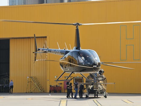 A helicopter stationed in front of a yellow hangar with people preparing for flight.