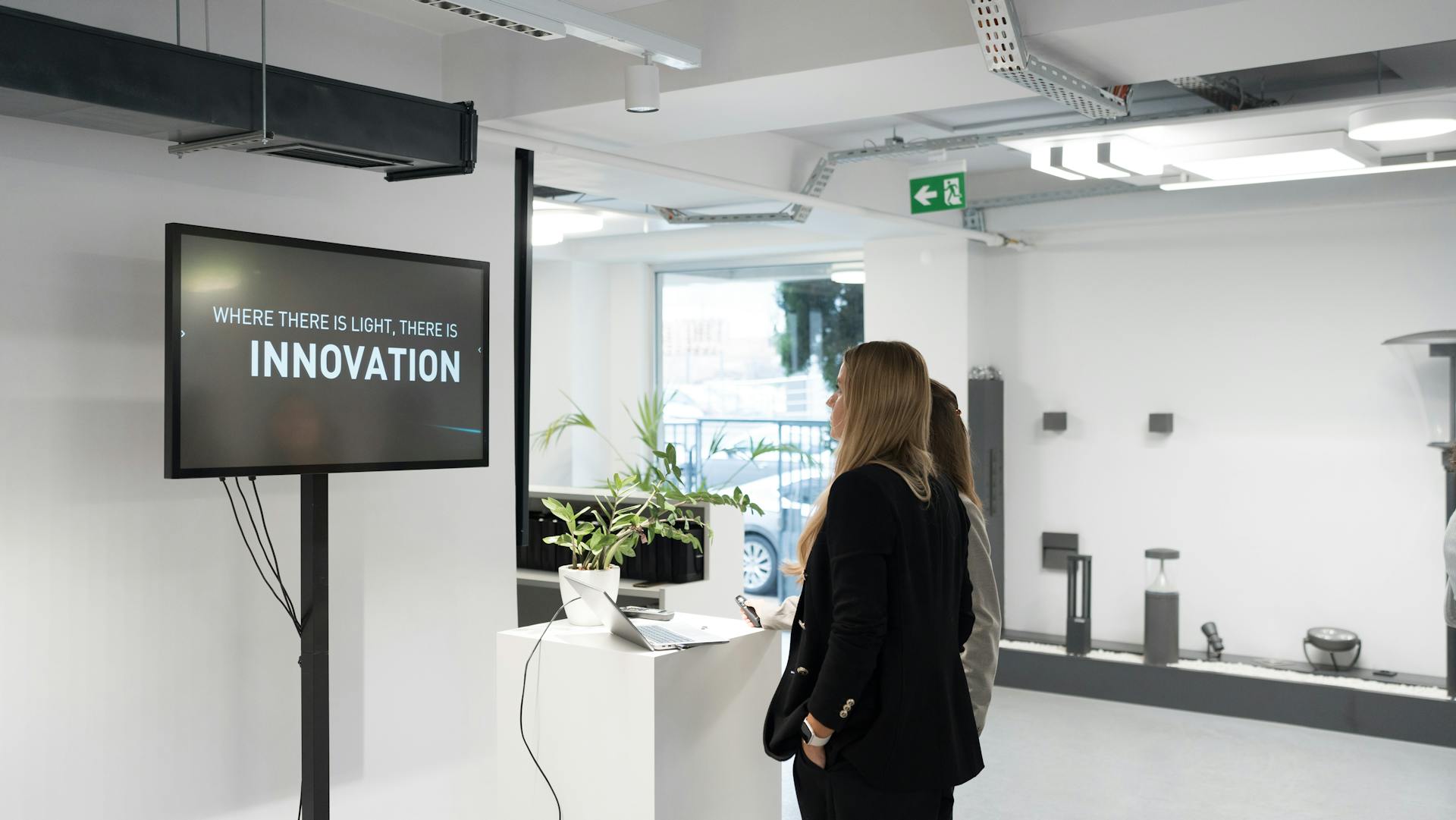 Two businesswomen discussing innovation in a modern office with a motivational screen.