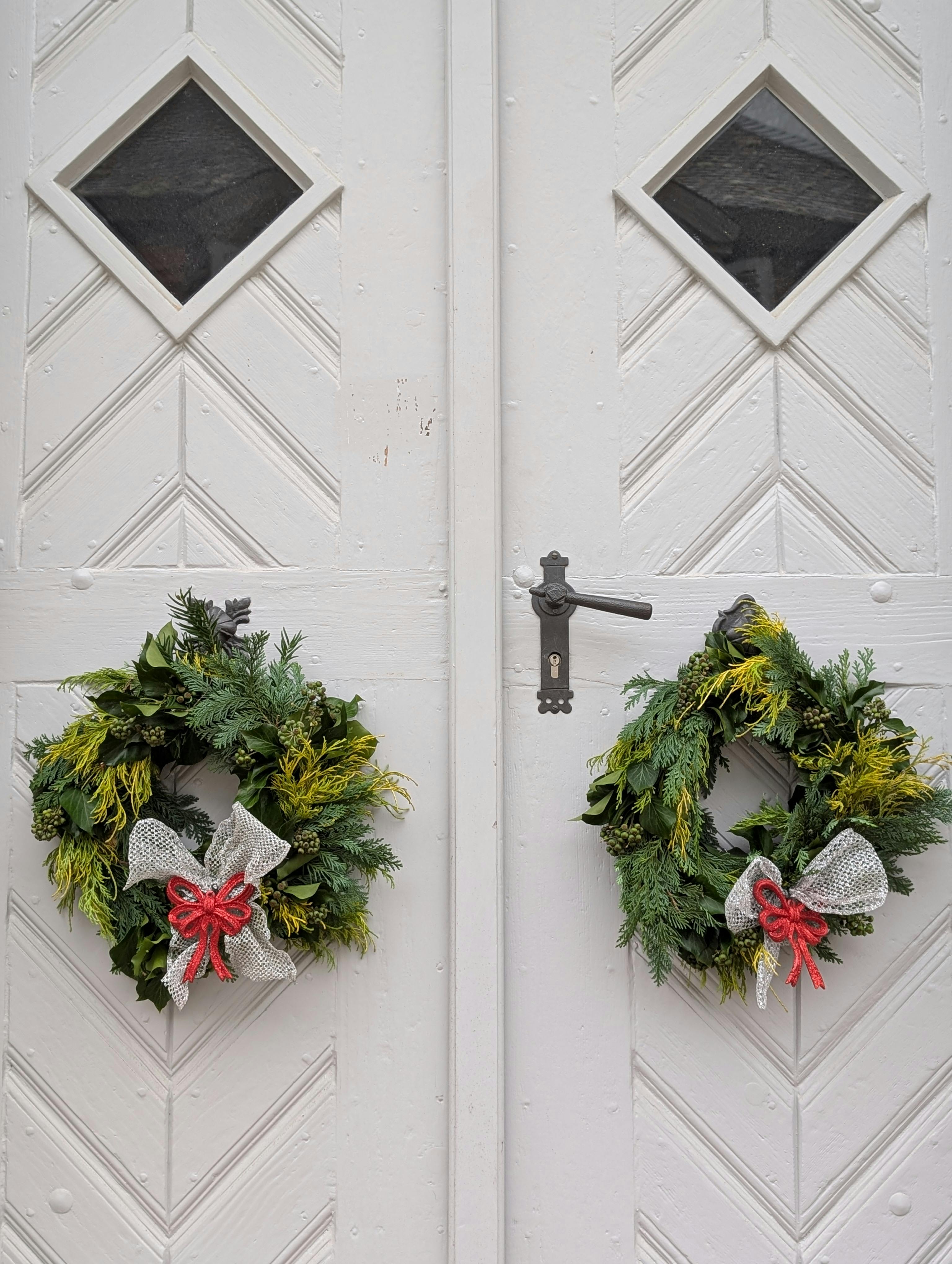 Elegant evergreen wreaths with red bows on white doors in a winter setting.