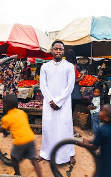 Man in white attire stands amidst vibrant market scene with colorful stalls and shoppers.