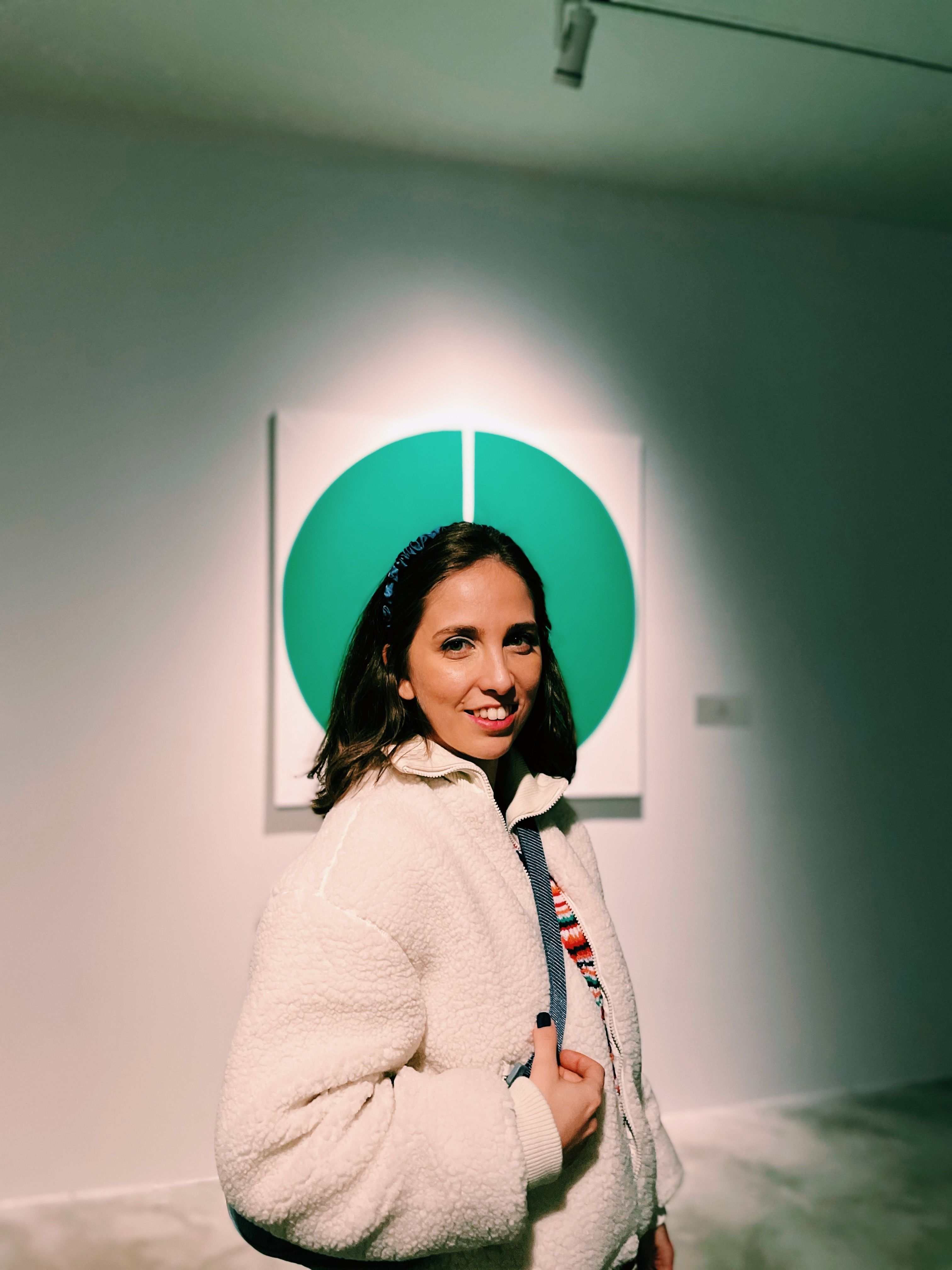 Gratis Una giovane donna con una giacca comoda si trova in una galleria d'arte con opere d'arte moderne. Foto a disposizione