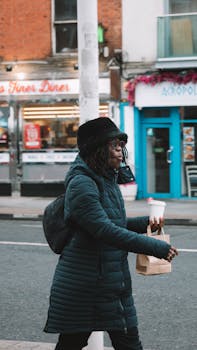 Woman in a winter coat walking with coffee and food bag in an urban setting.
