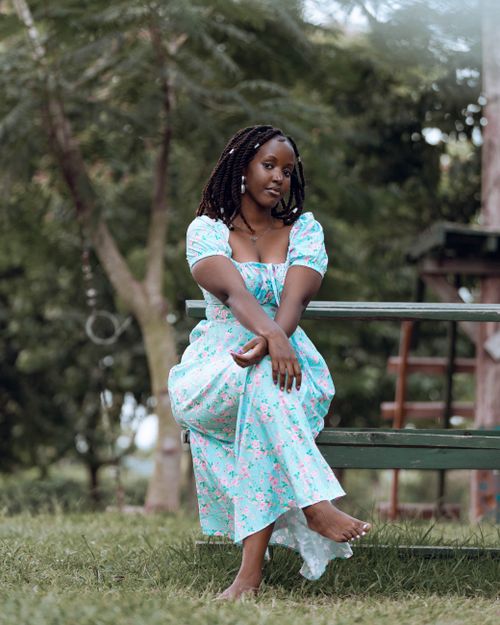 Free A young woman in a floral dress sitting on a bench in a lush green park, enjoying a peaceful day outdoors. Stock Photo