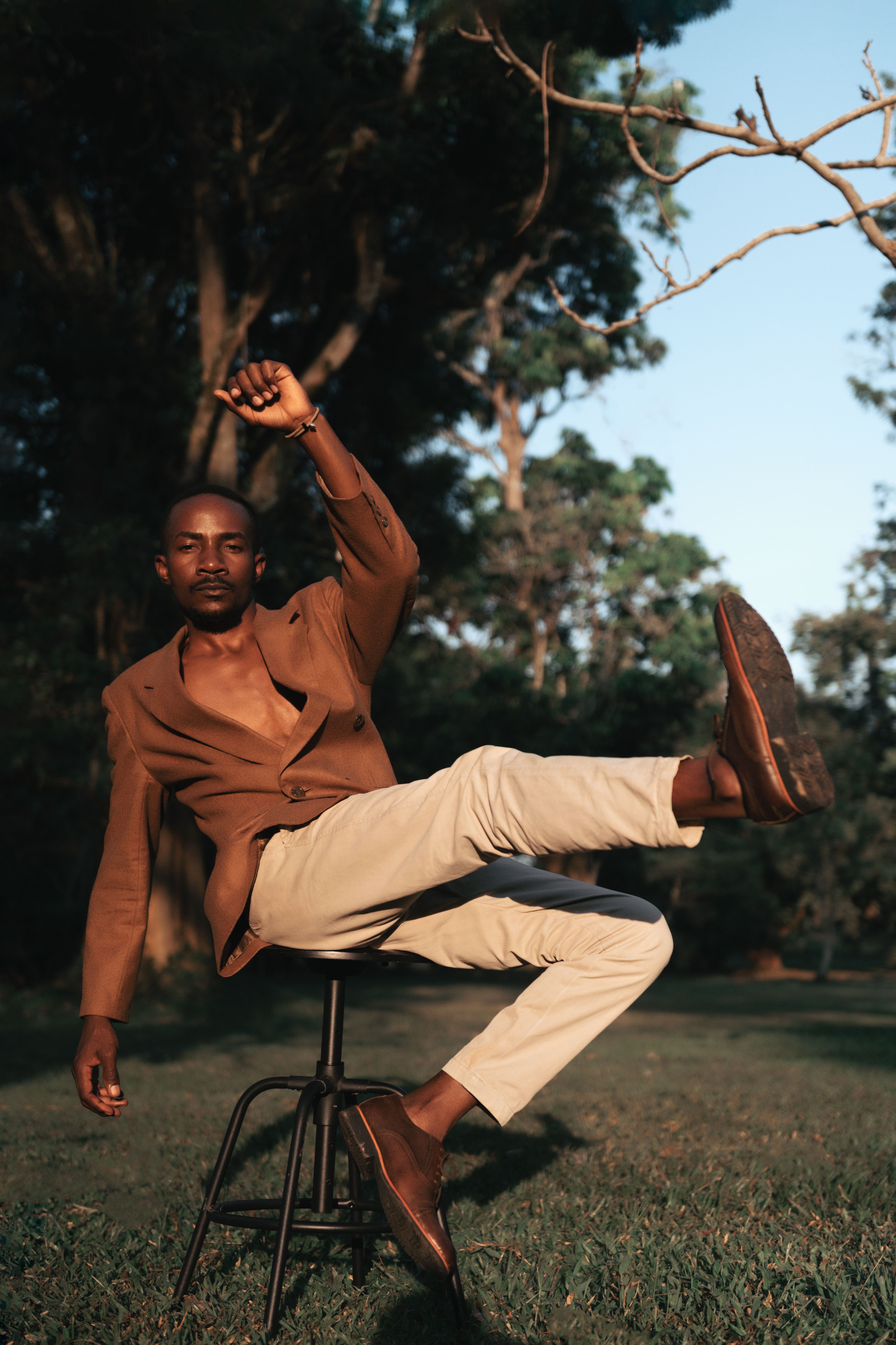 A fashionable man in a brown blazer strikes a dynamic pose on a chair in a sunlit park.