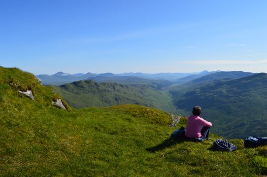 A woman sits on a grassy hilltop, admiring the stunning Scottish Highlands under a clear blue sky.