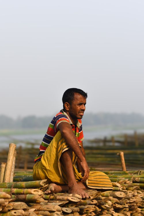 Free Man sitting on bamboo near a riverbank in Narayanganj, showcasing traditional life in Bangladesh. Stock Photo