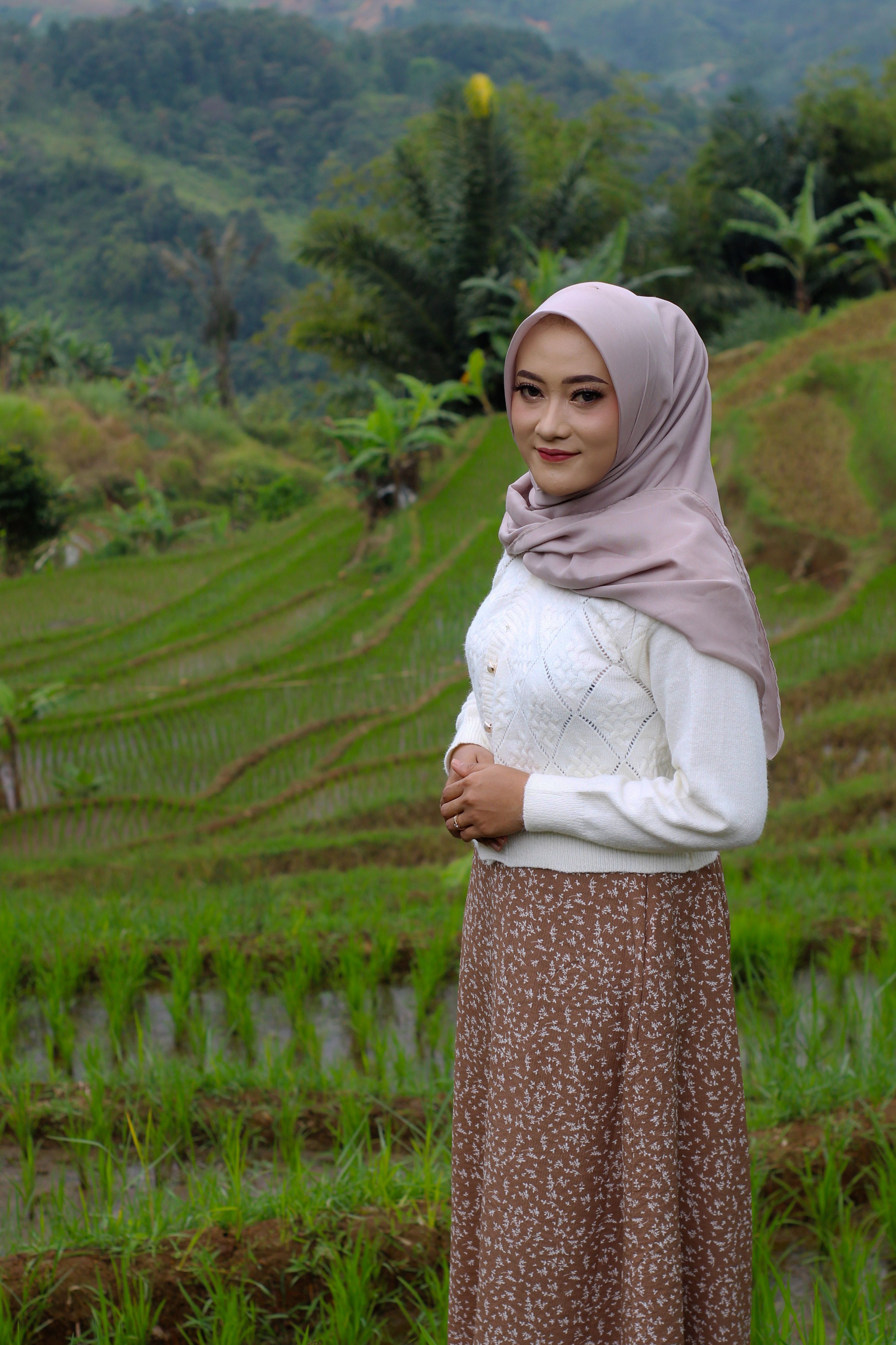 A woman in a hijab stands in a vibrant rice field with lush green terraces.