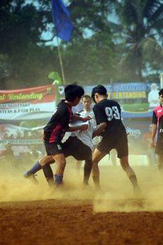 A dynamic football match with teenagers playing on a dusty outdoor field.