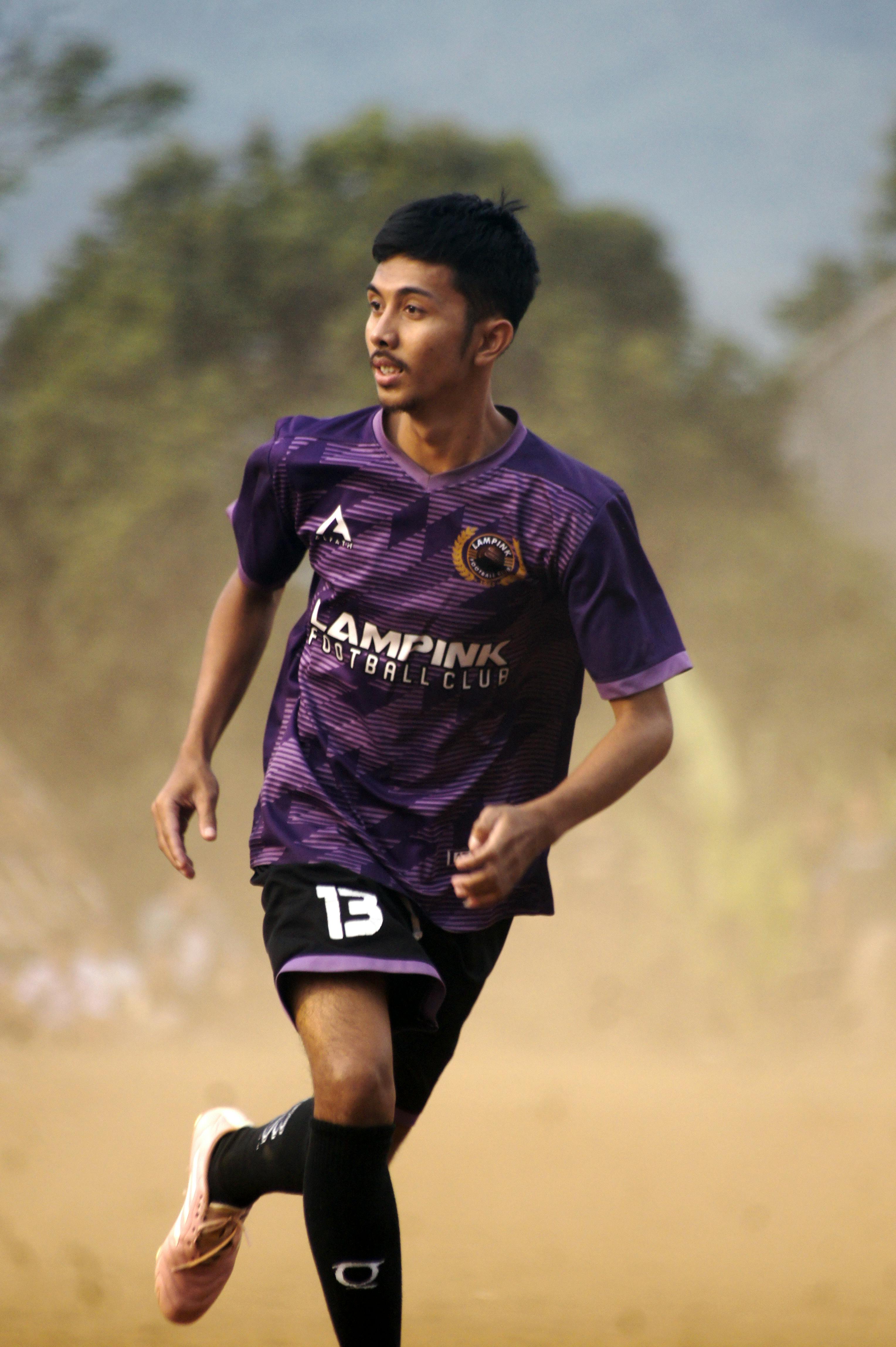 A football player in motion on a dusty field, wearing a purple jersey during daytime match.