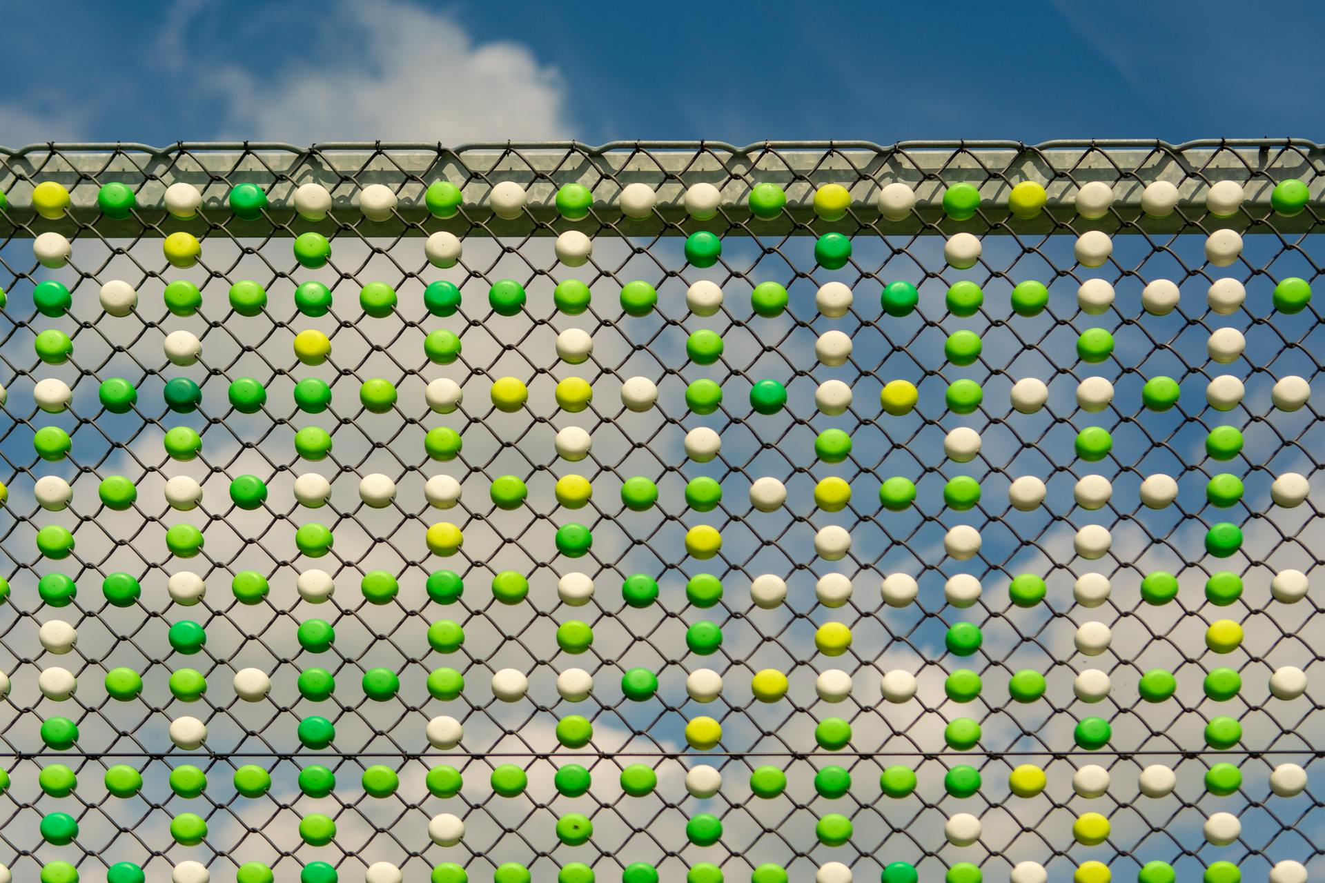 Colorful beads in a pattern on a chainlink fence contrast with a blue sky and clouds.