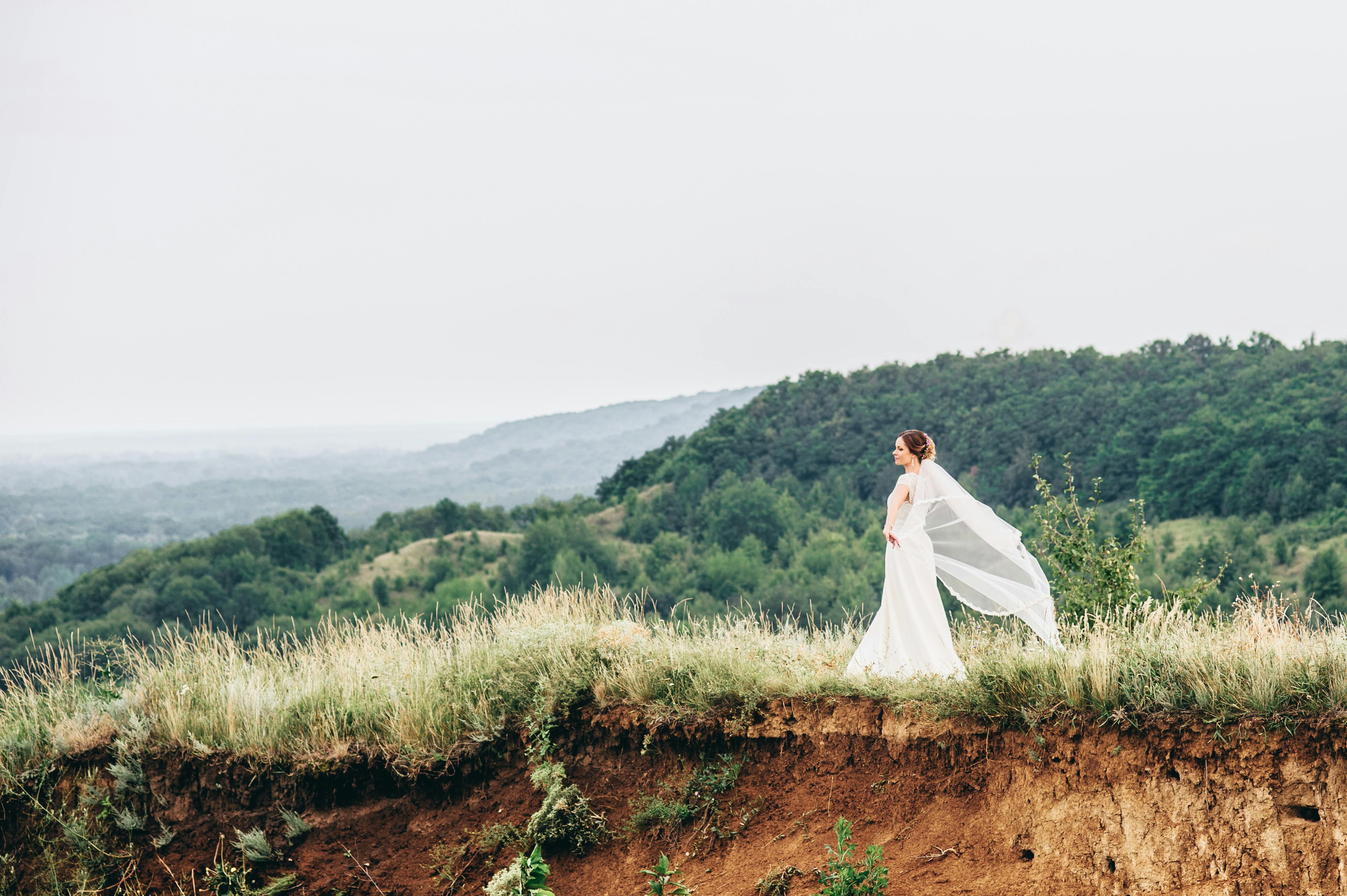 Bride in white dress on a cliff with flowing veil and lush landscape backdrop.