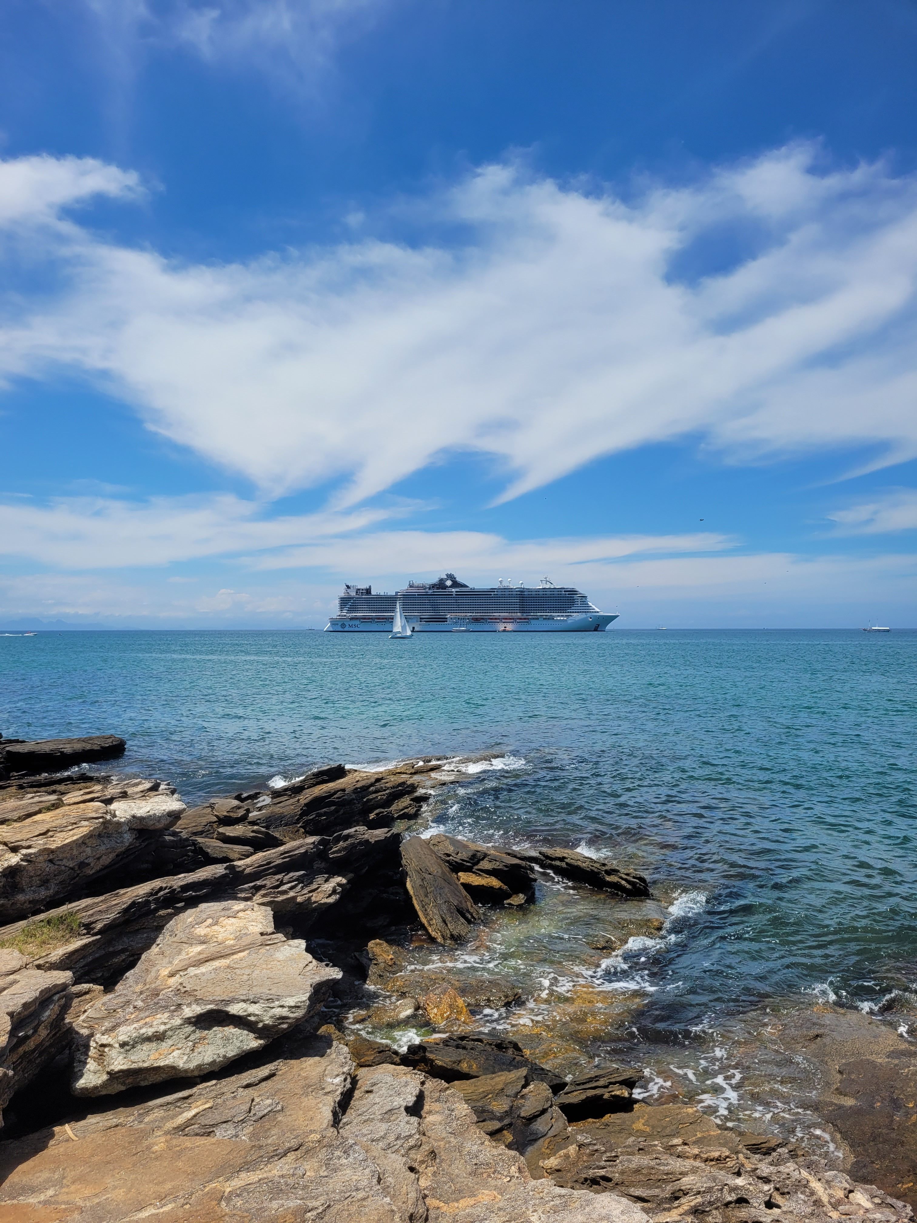 Cruise ship sailing on a clear blue ocean day with rocky shore and picturesque sky.