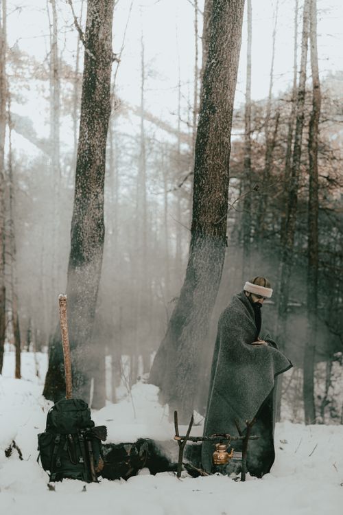 Free A person wrapped in a blanket standing by a campfire in a snowy forest. Stock Photo