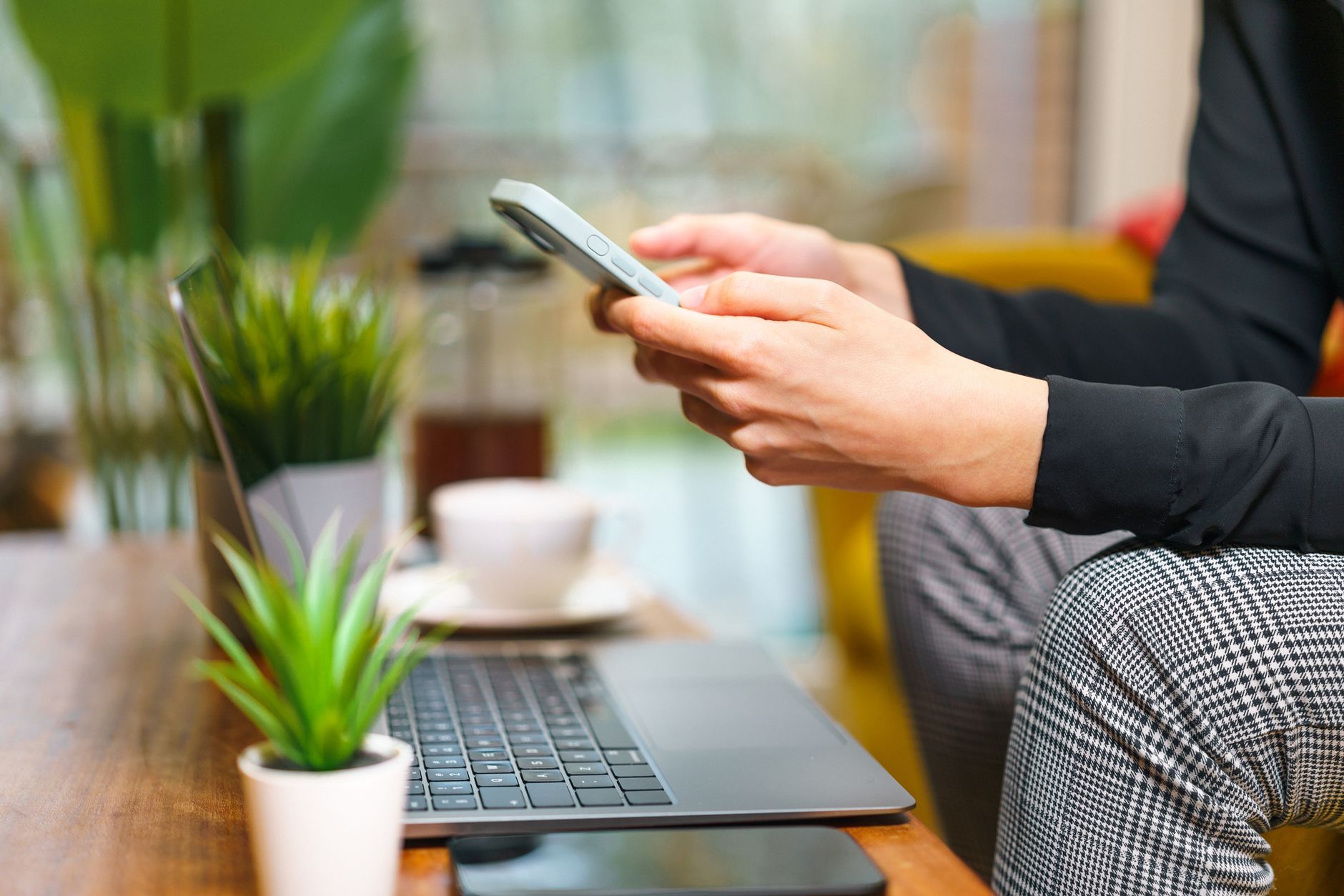 A person using a smartphone in a cozy home office setting with plants, a laptop, and coffee.