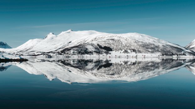 Captivating arctic landscape with snow-covered mountains mirrored in a serene lake under a bright sky.
