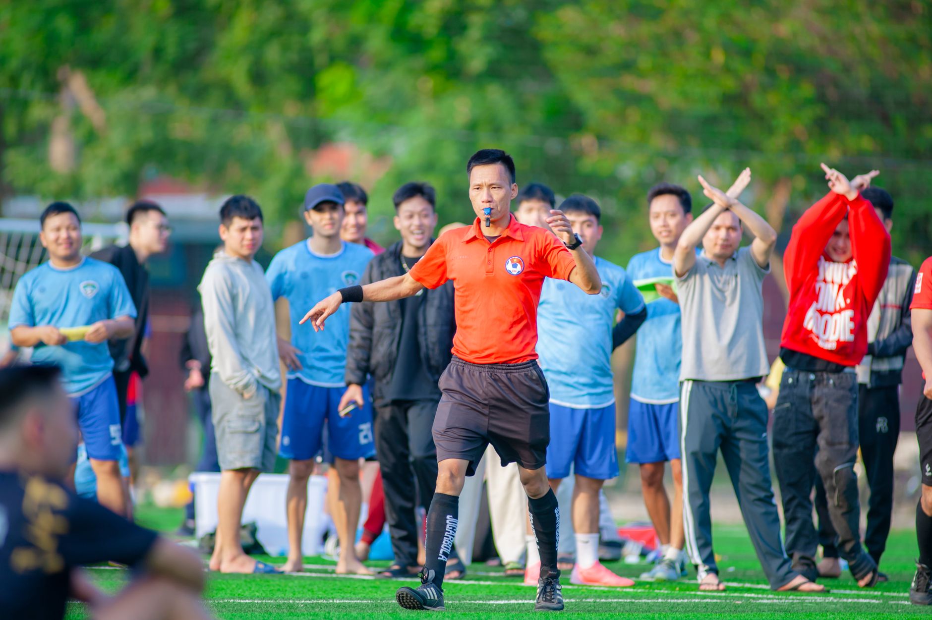 A referee in orange signals during a soccer game with players watching in the background.