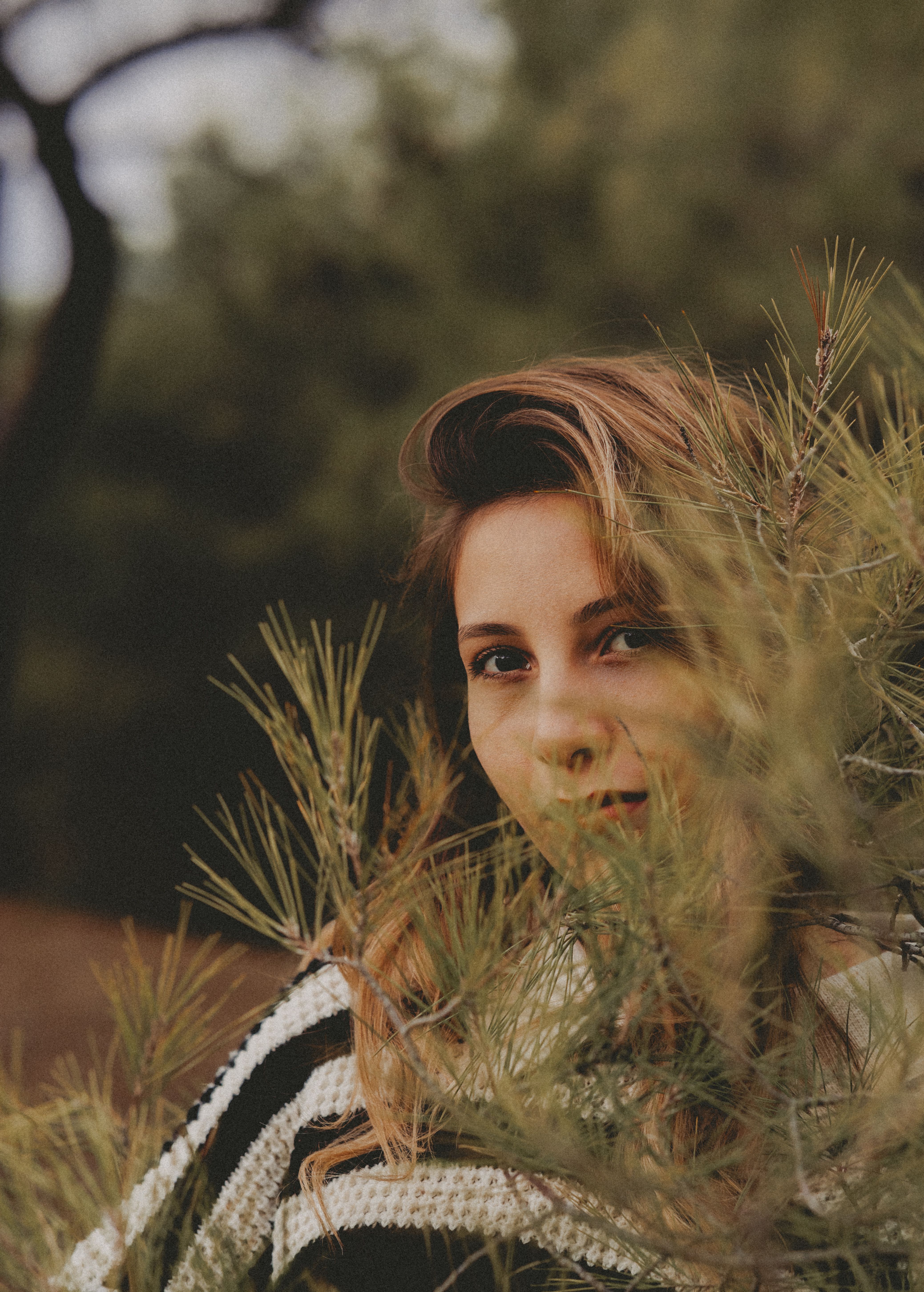 Young woman peering through pine branches, outdoor portrait with natural surroundings.