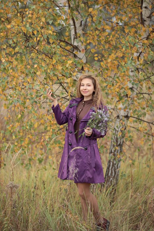 Free A young woman in a purple coat stands holding a small bouquet in front of autumnal trees. Stock Photo