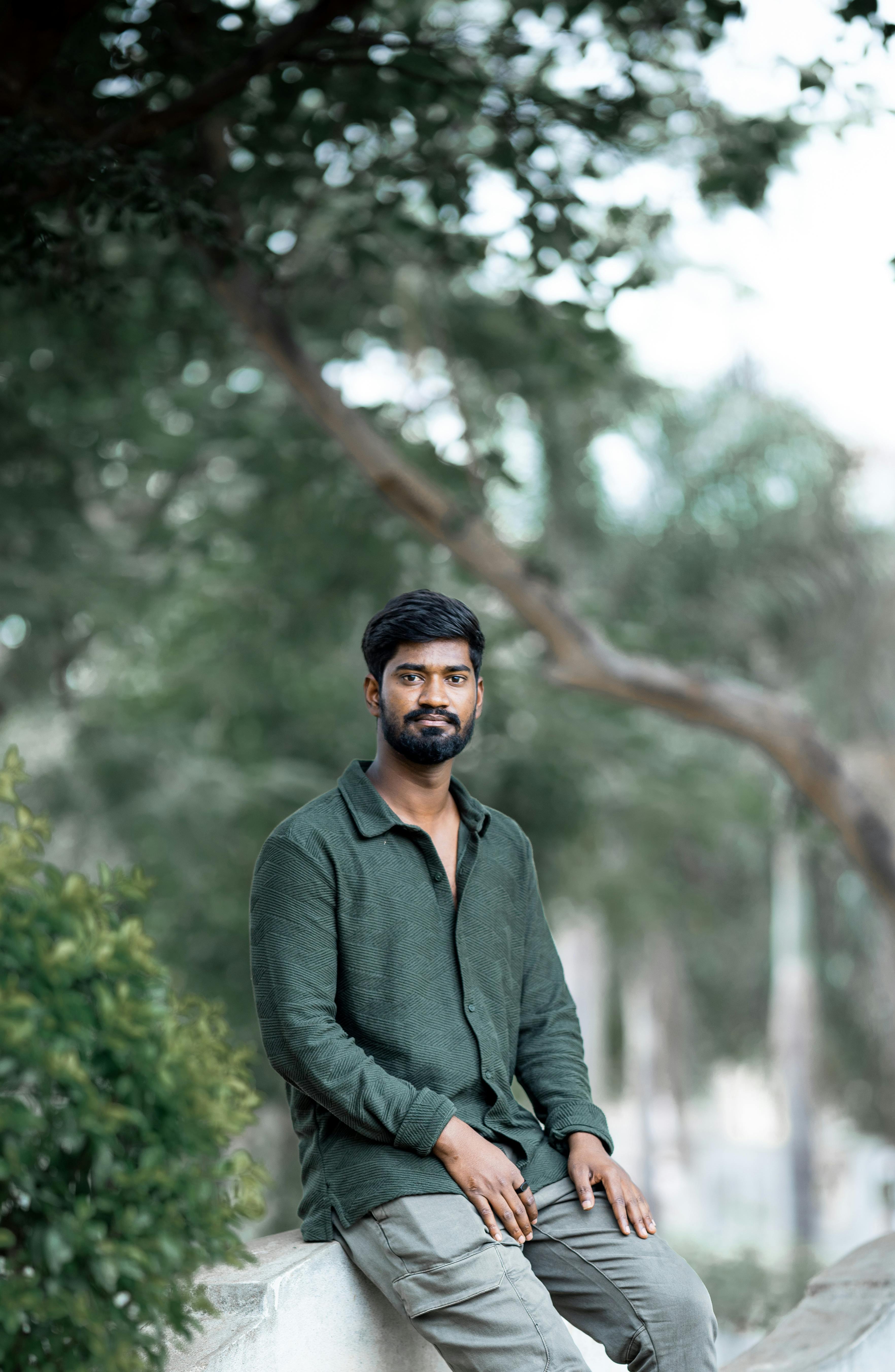 A young man in a green shirt sitting on a stone, surrounded by greenery in an outdoor setting.
