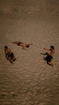Three people interact playfully on a sandy beach viewed from above.
