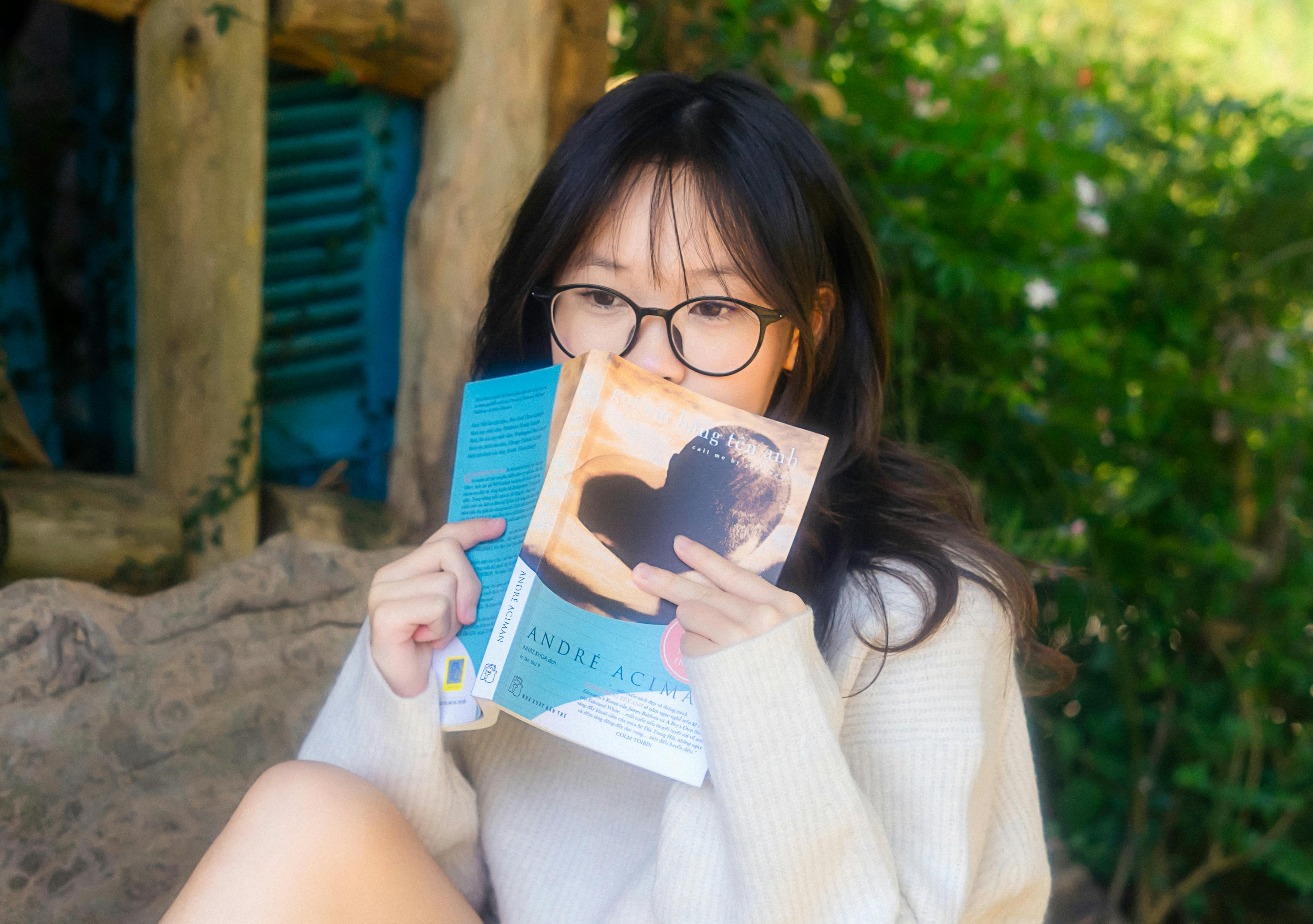 A young woman with glasses enjoys reading a book in a serene outdoor garden.
