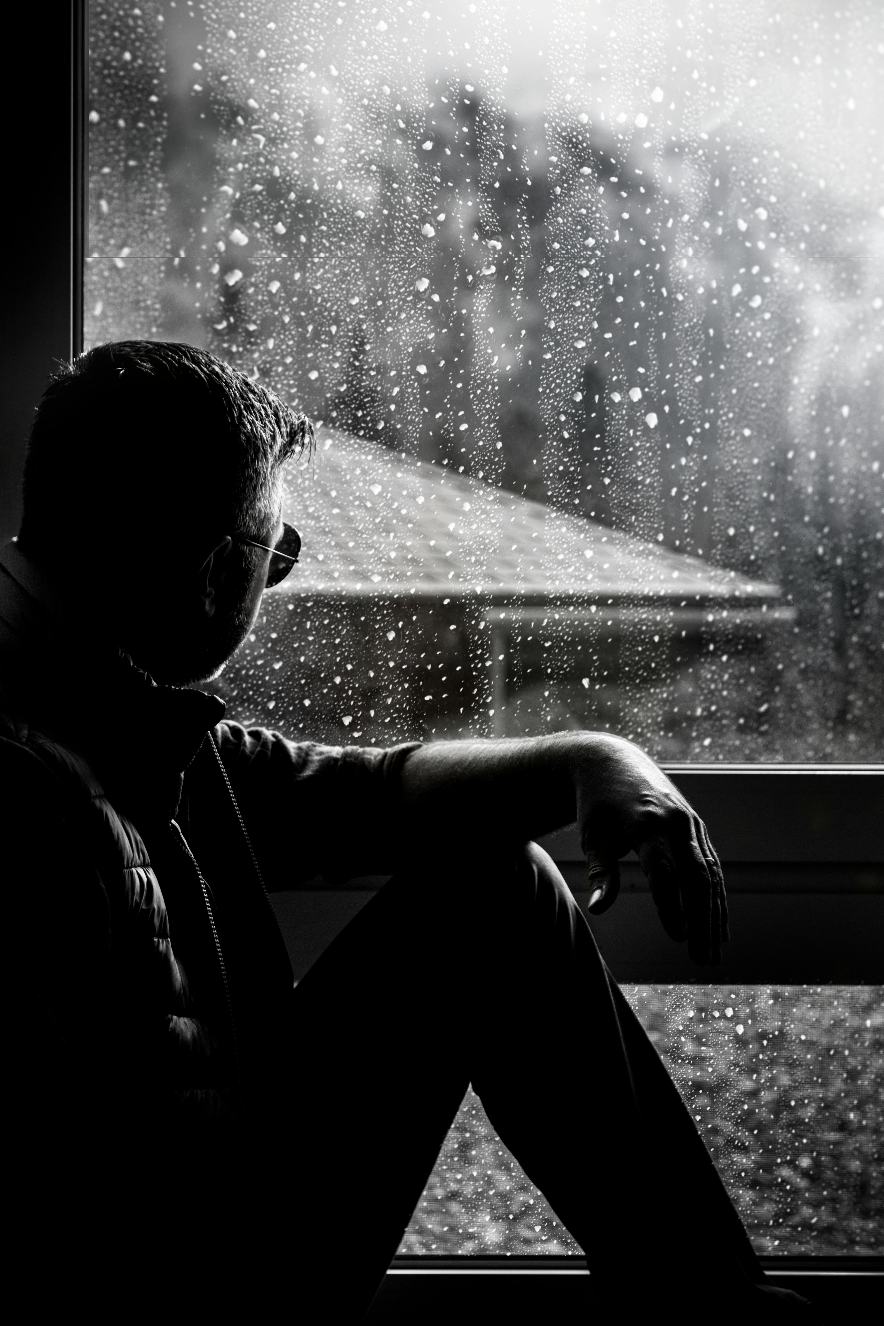 grayscale photo of man sitting beside window